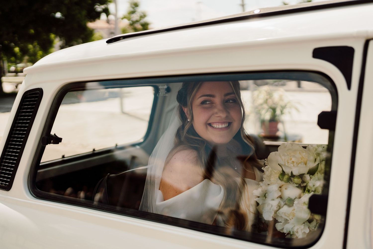 Smiling bride holding white bouquet looking out the window of a vintage white vehicle.