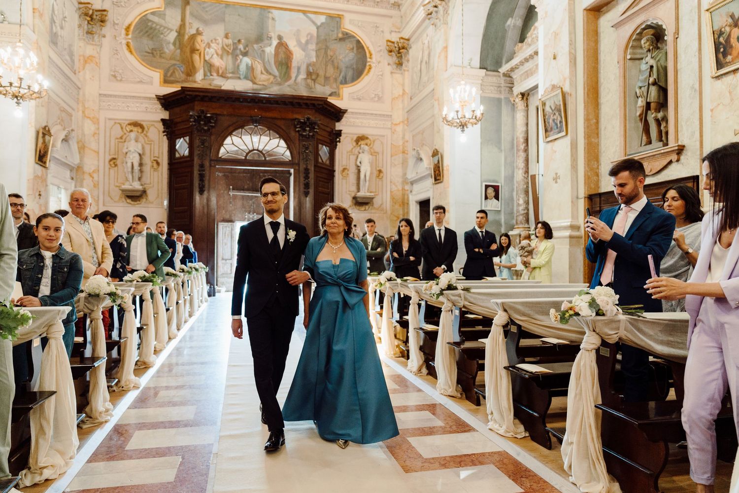 Groom escorting a woman in teal down the aisle of an ornate Italian church wedding.