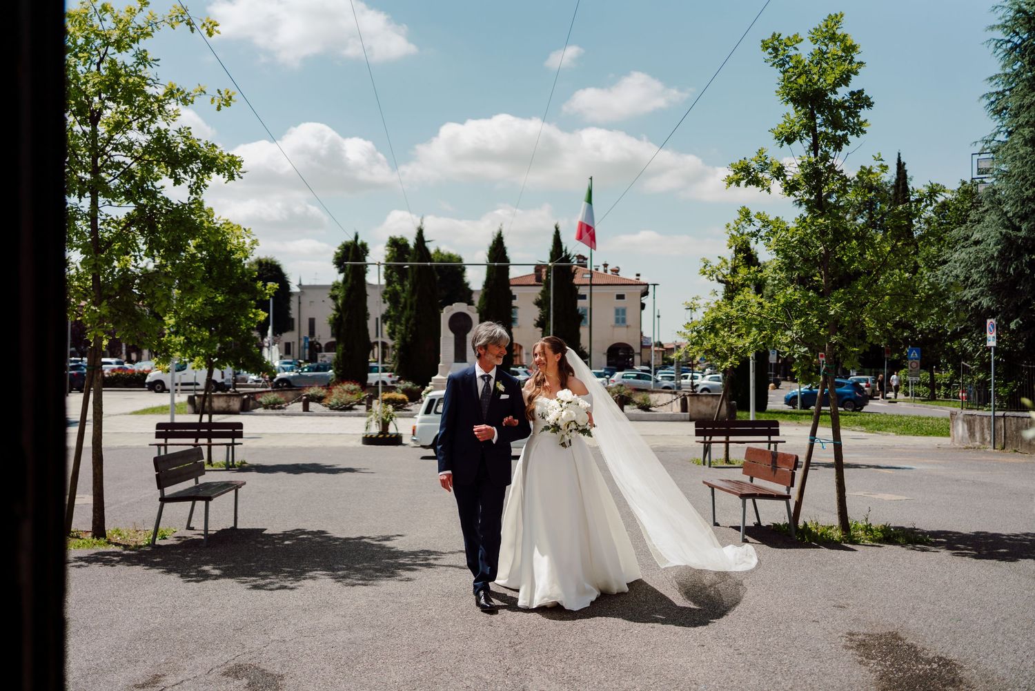 Bride in a ball gown walking with her father outdoors on a sunny wedding day.