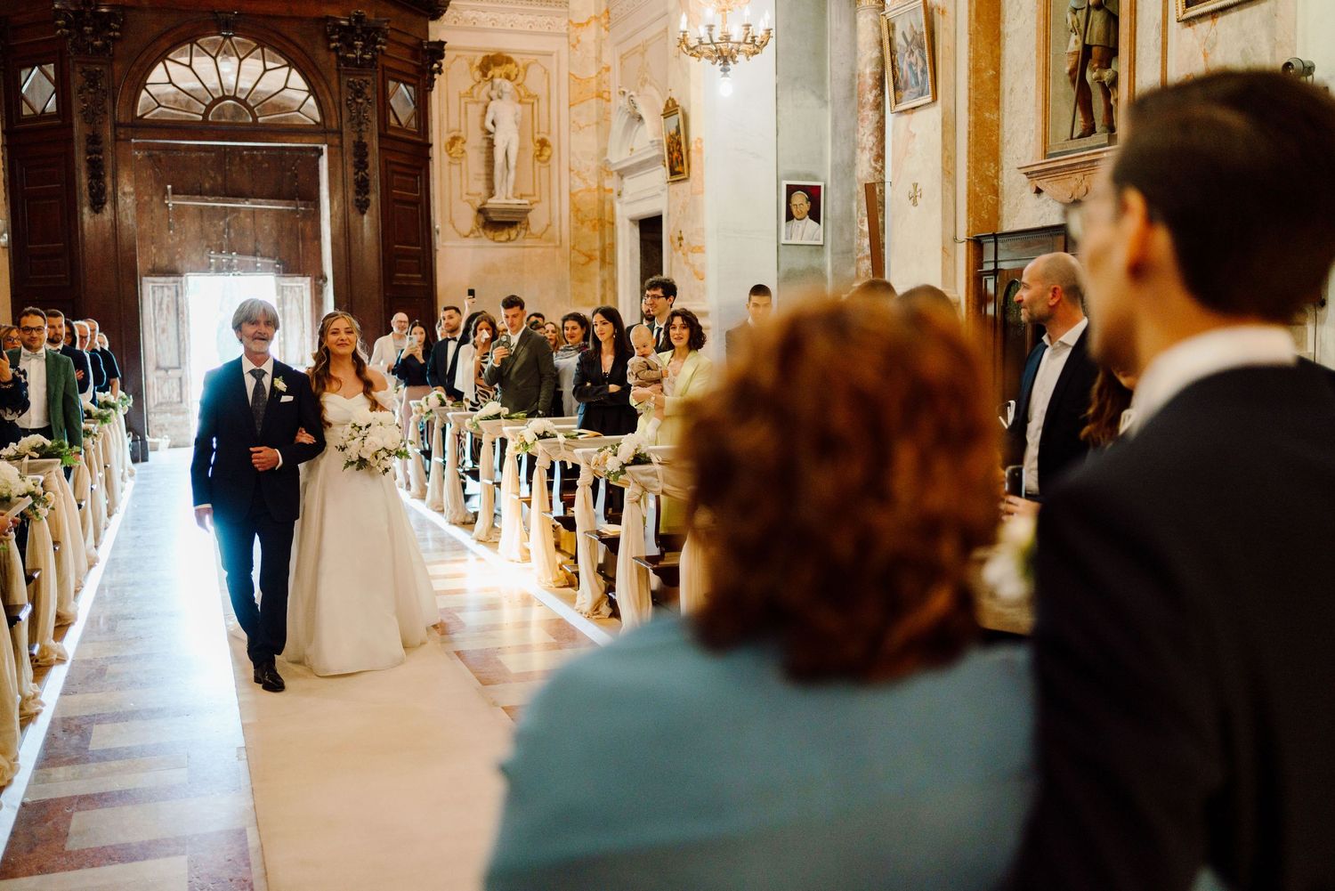 Bride walking down the aisle with her escort in a beautifully decorated baroque church.