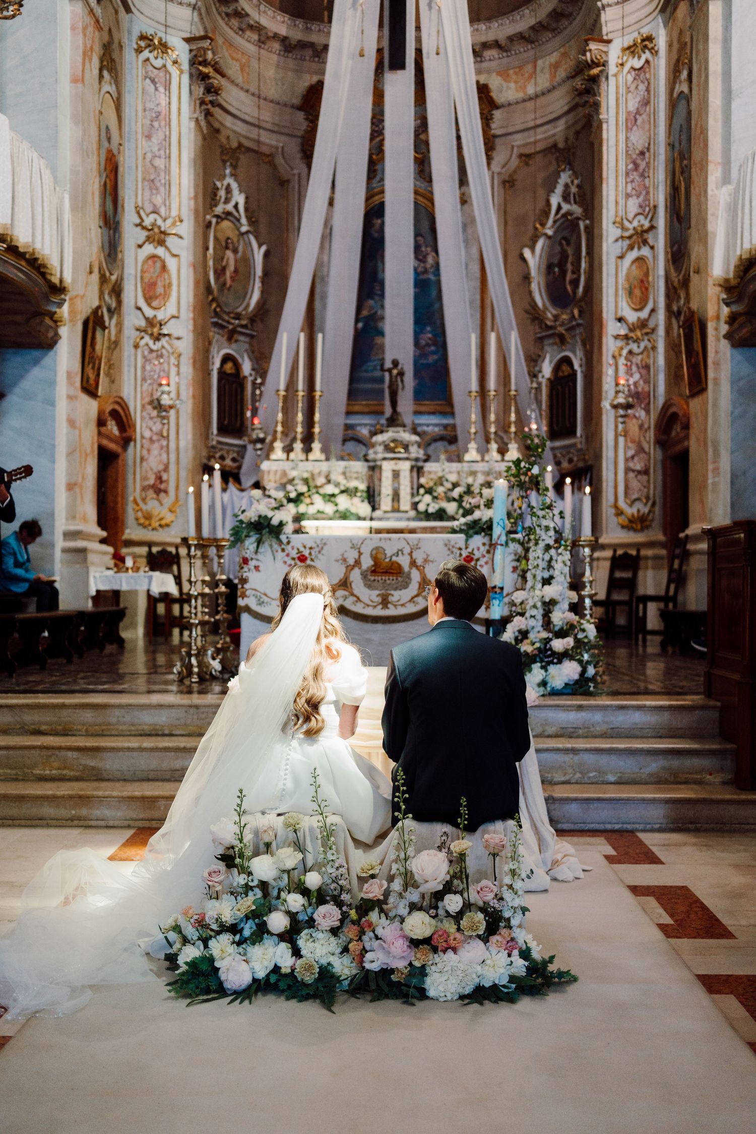Bride and groom kneel before ornate baroque church altar surrounded by floral arrangements.