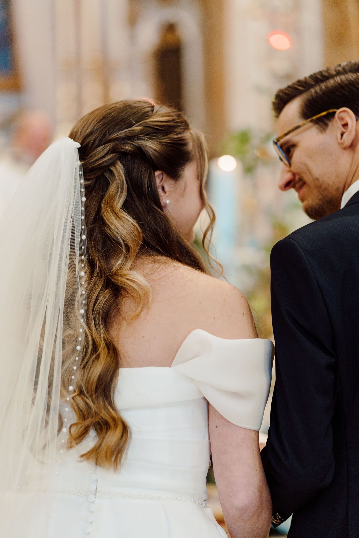 Close-up of brides elegant off-shoulder gown and pearl-adorned veil in church.
