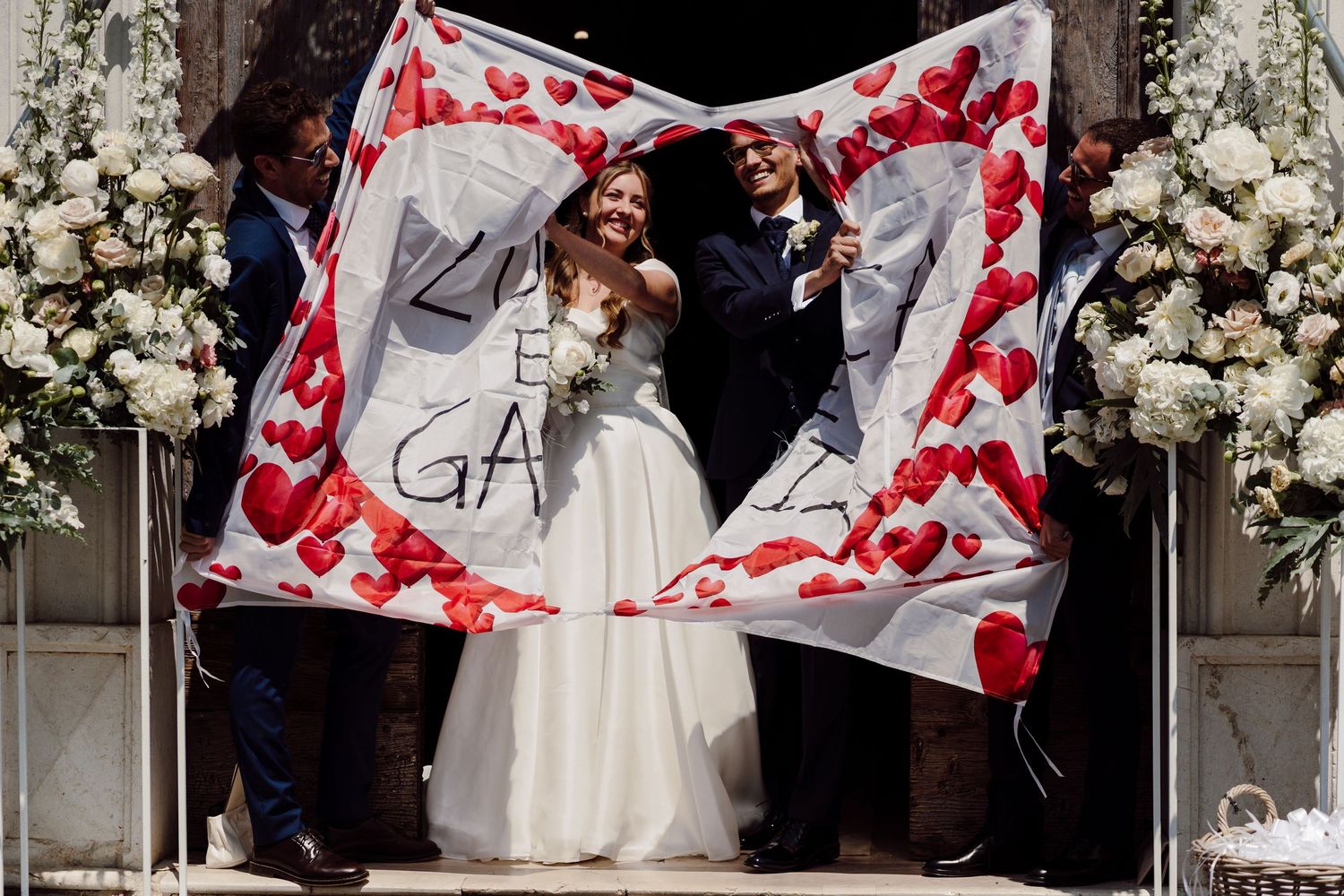 Newlyweds emerge from church entrance under a festive banner decorated with red hearts.