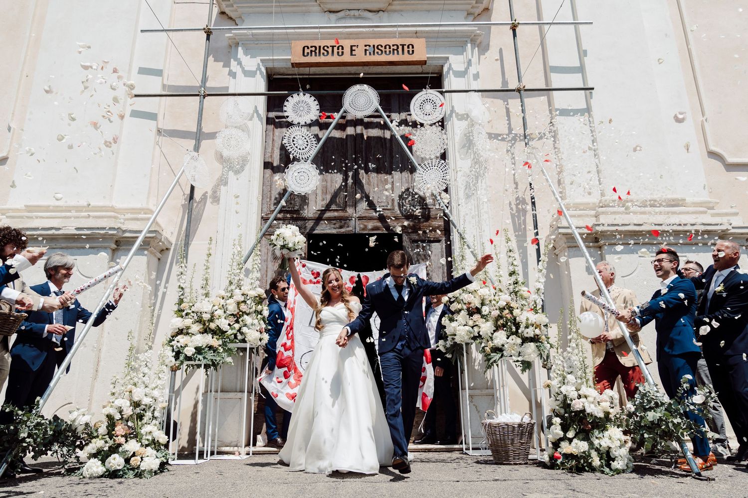 Bride and groom exit church doors as guests celebrate with confetti at a sunny Italian wedding ceremony.