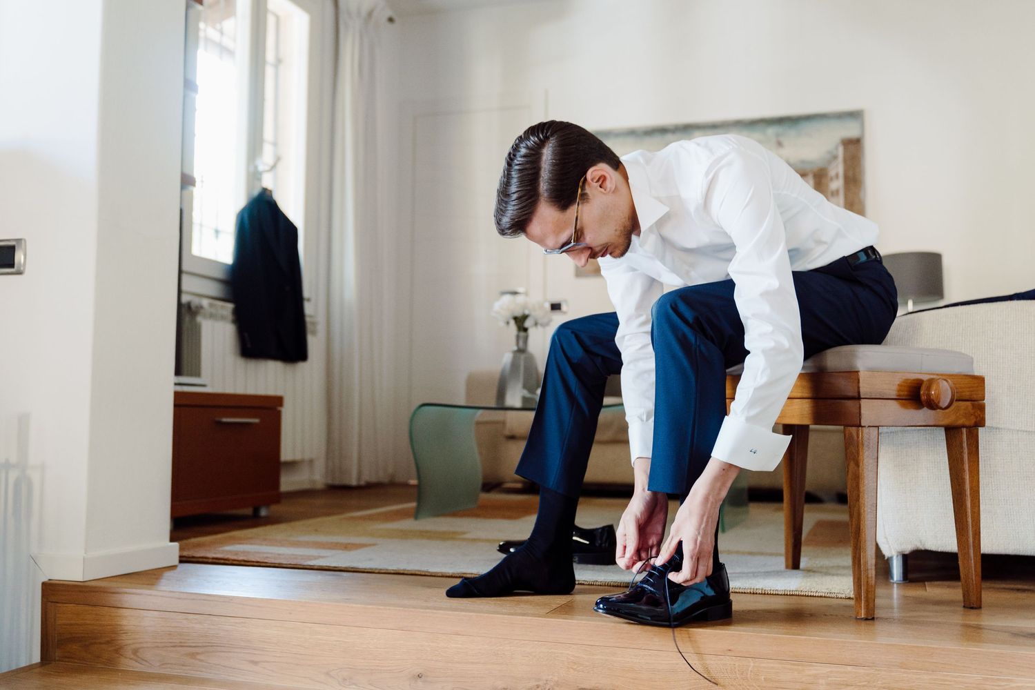 Groom in white shirt and blue trousers tying his dress shoes in a bright apartment.