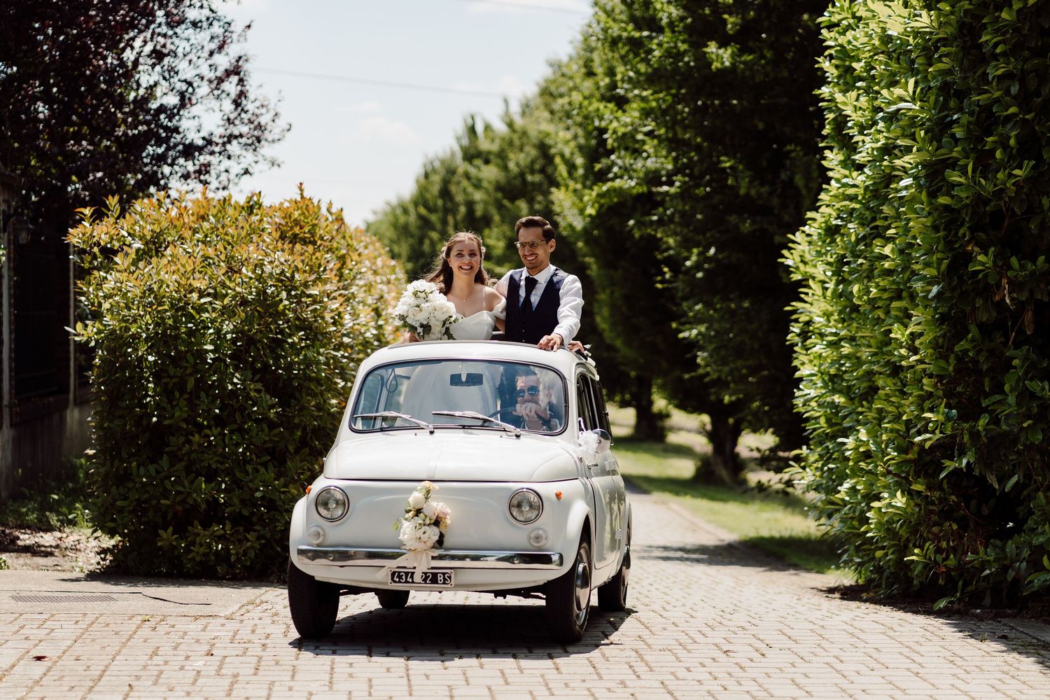 Newlyweds ride in a classic white Fiat 500 vintage car through a tree-lined driveway on their wedding day.