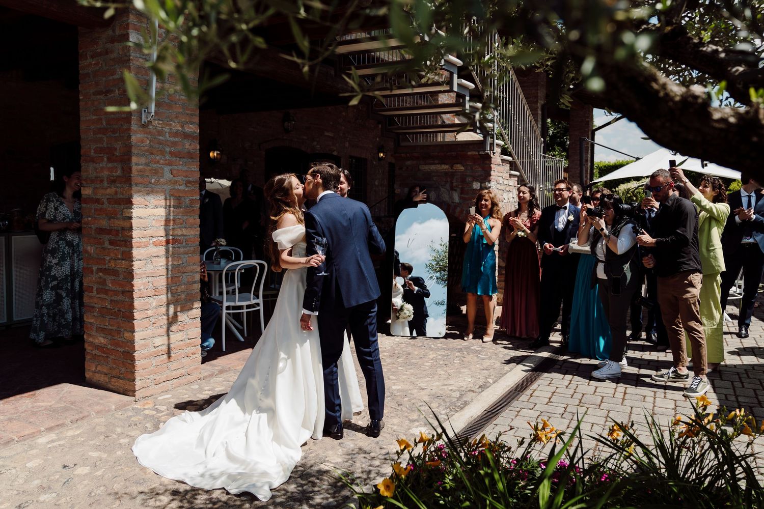 Bride and groom share a kiss while wedding guests applaud at an outdoor Italian villa reception.