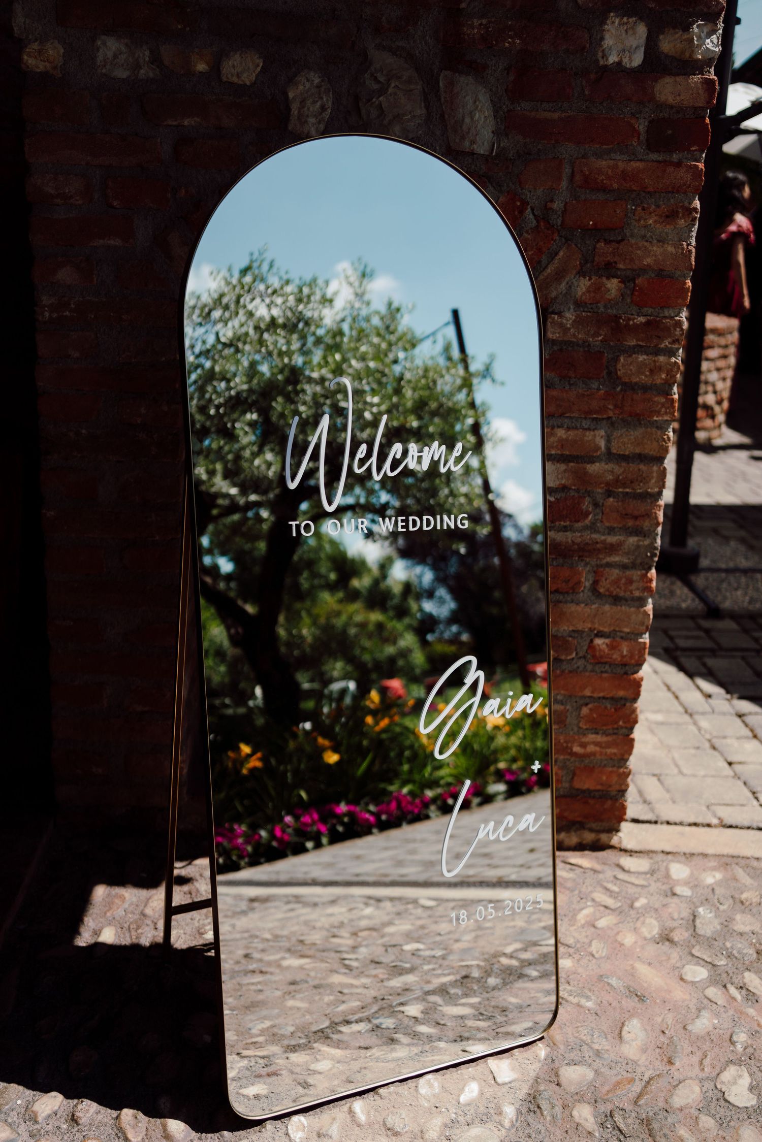 Elegant arched mirror wedding welcome sign with white script lettering leaning against a rustic brick wall.