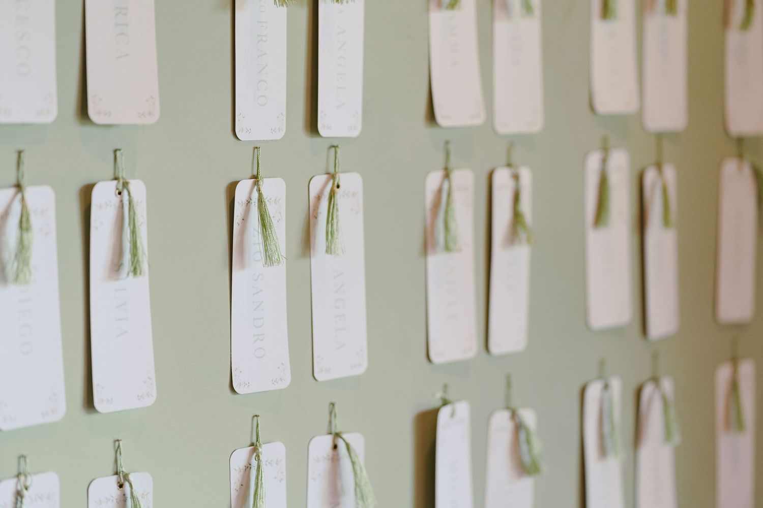 White wedding escort cards with sage green ribbons displayed on a soft green wall.