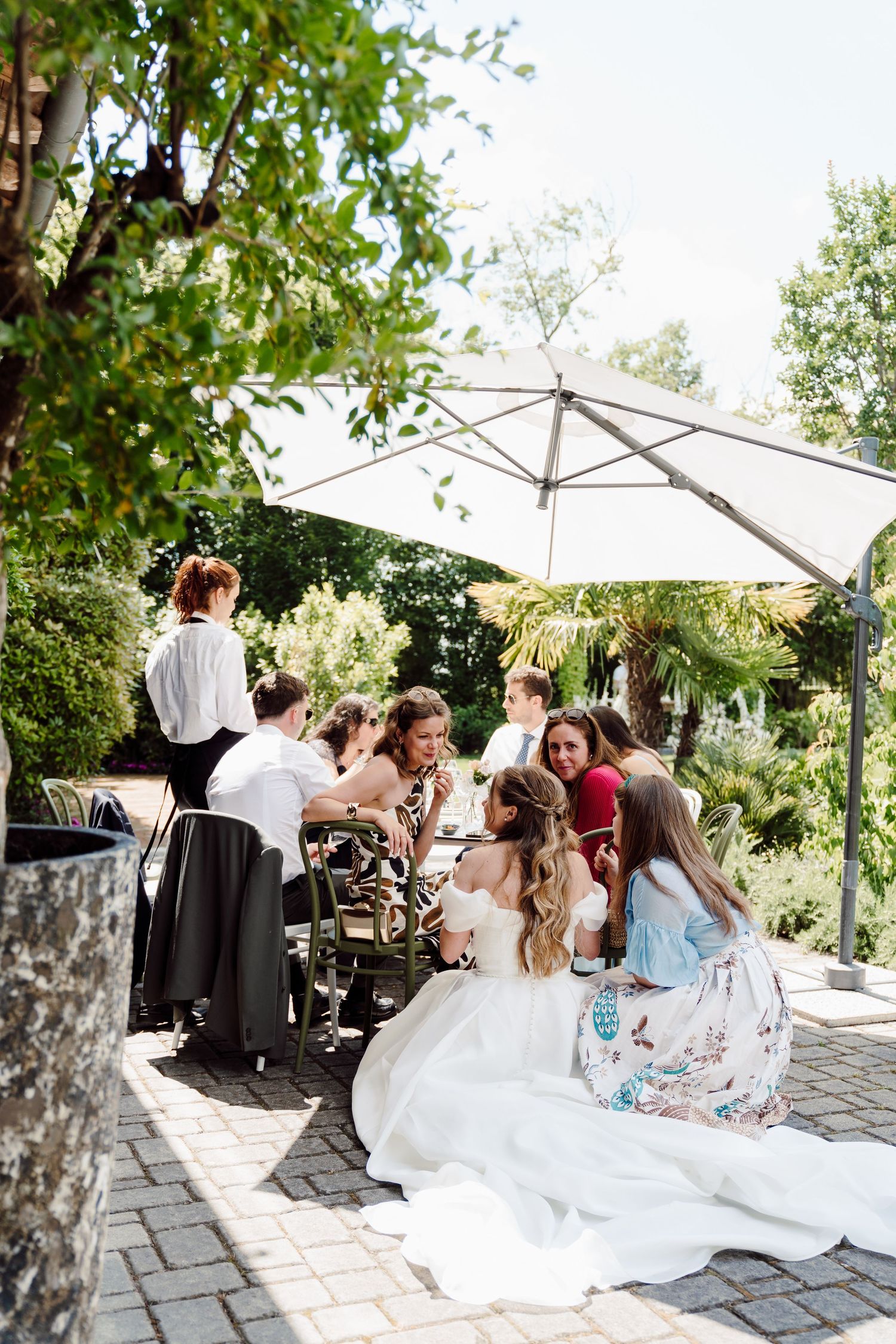 Bride in white gown sits with guests at an outdoor wedding reception table under a large umbrella.