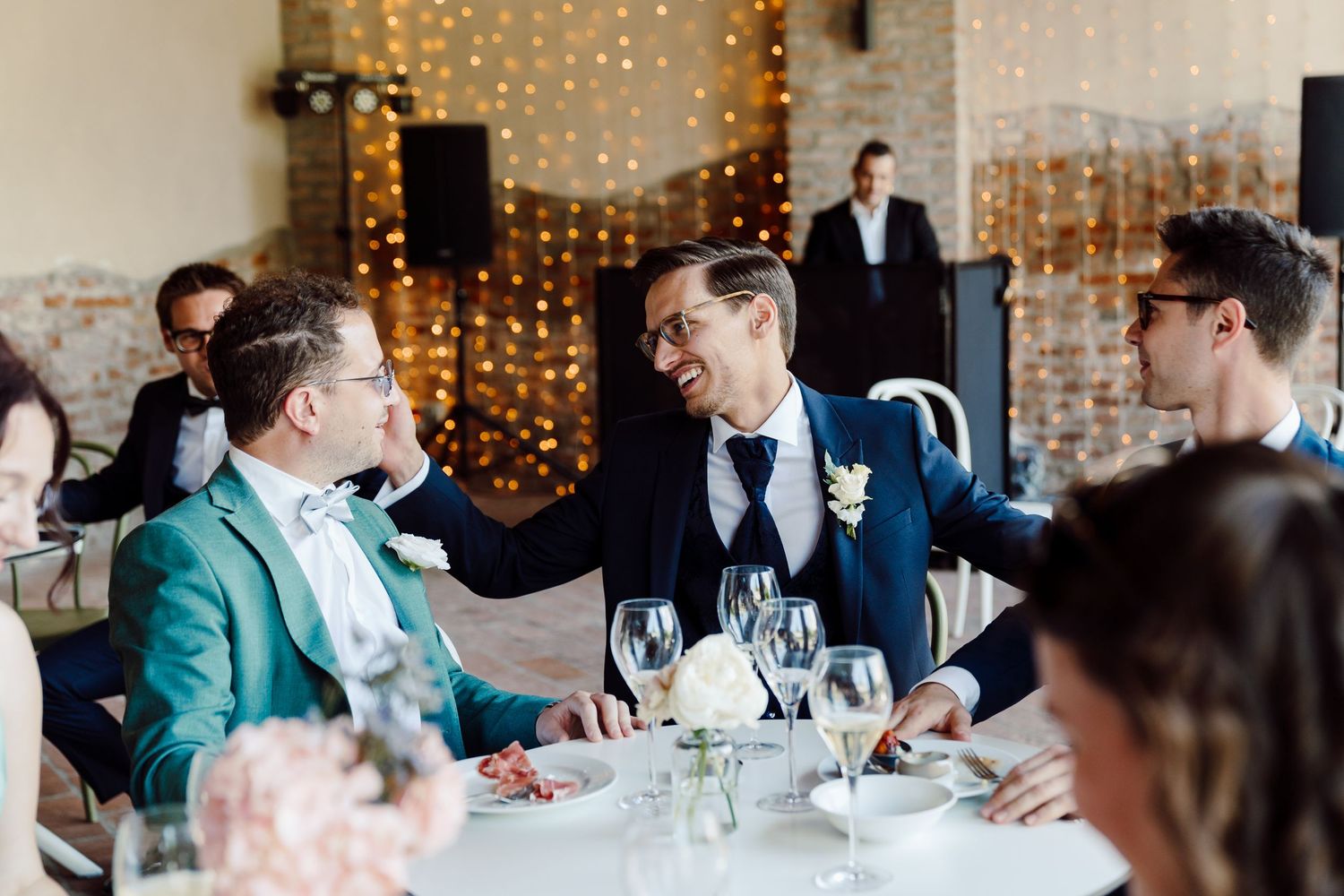 Groom in teal suit and man in navy suit share a joyful moment at elegant wedding reception dinner.