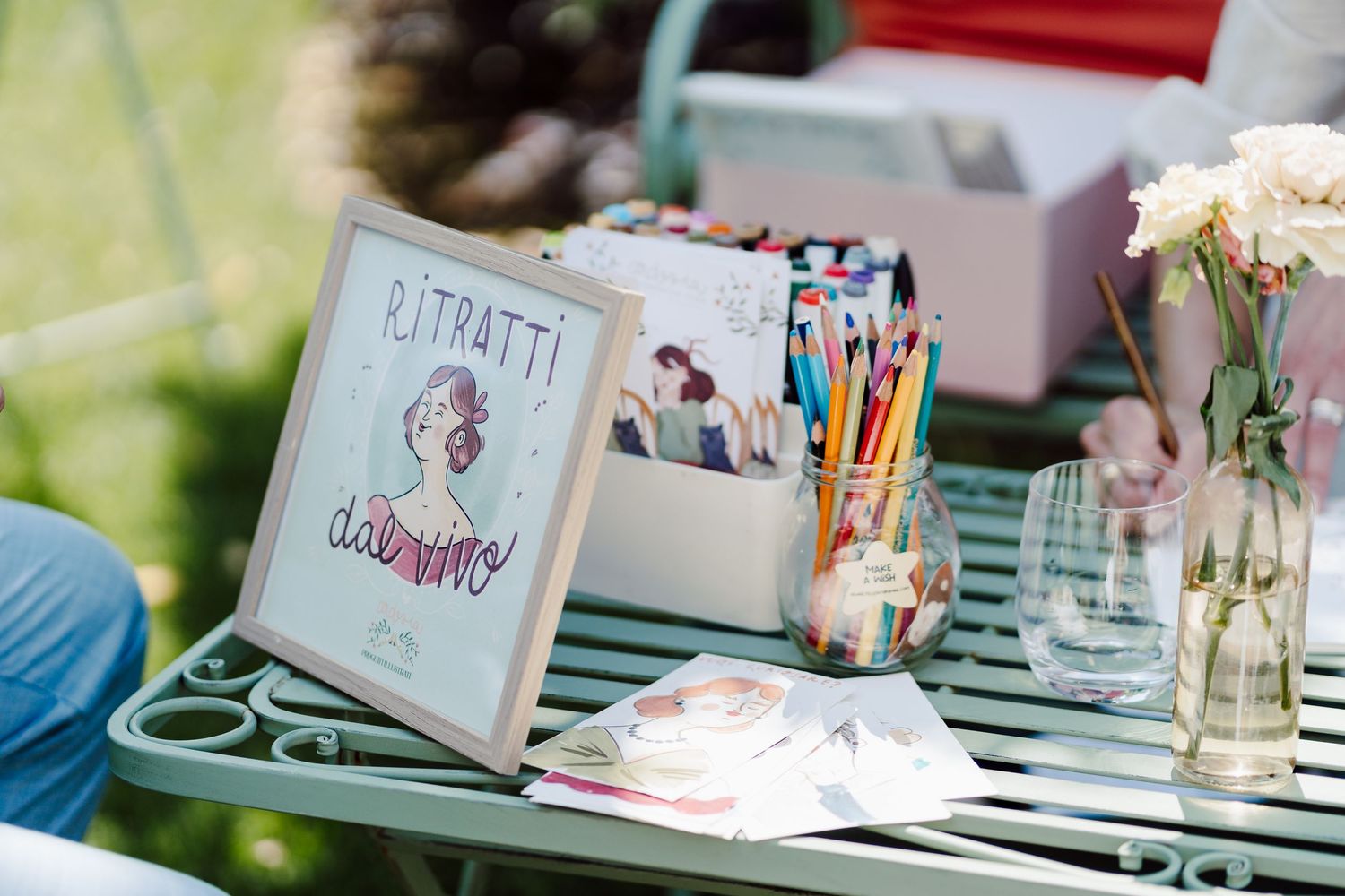 Wedding portrait station with illustrated sign, colored pencils, and floral decorations on a garden table.