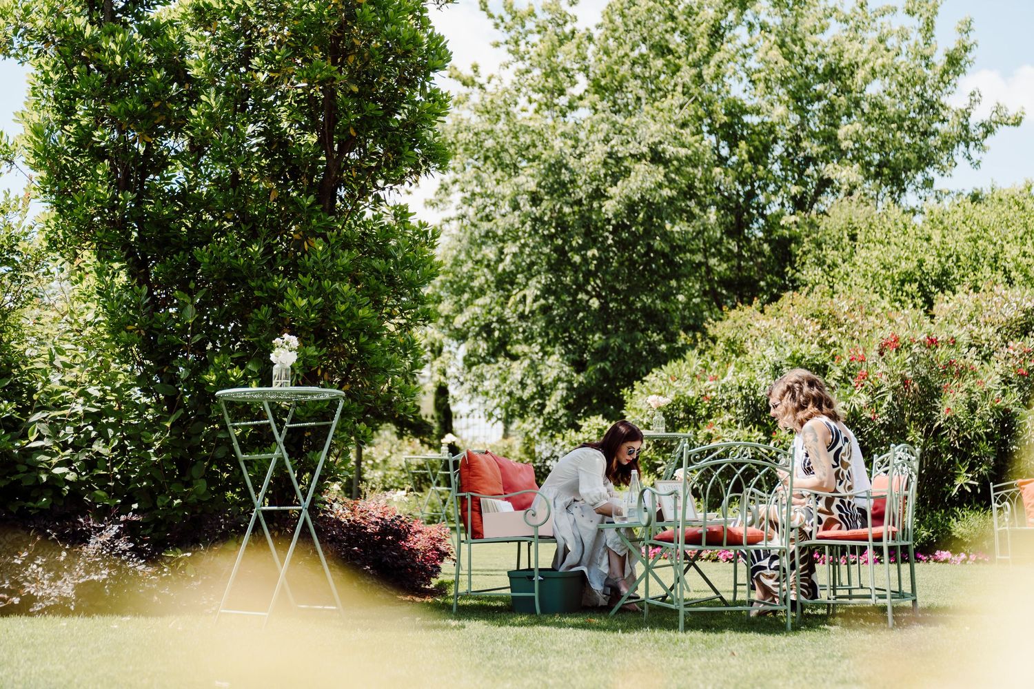 Two wedding guests enjoy conversation seated at decorated garden tables surrounded by lush green trees.