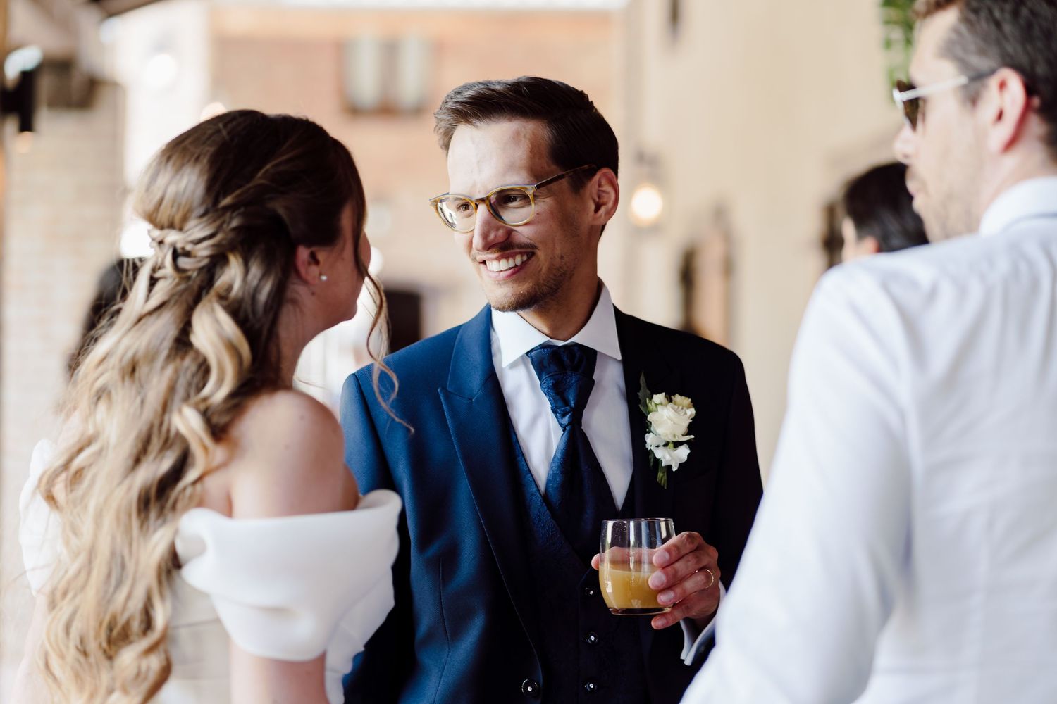 Smiling groom in navy suit holds champagne while chatting with bride at outdoor wedding cocktail hour.