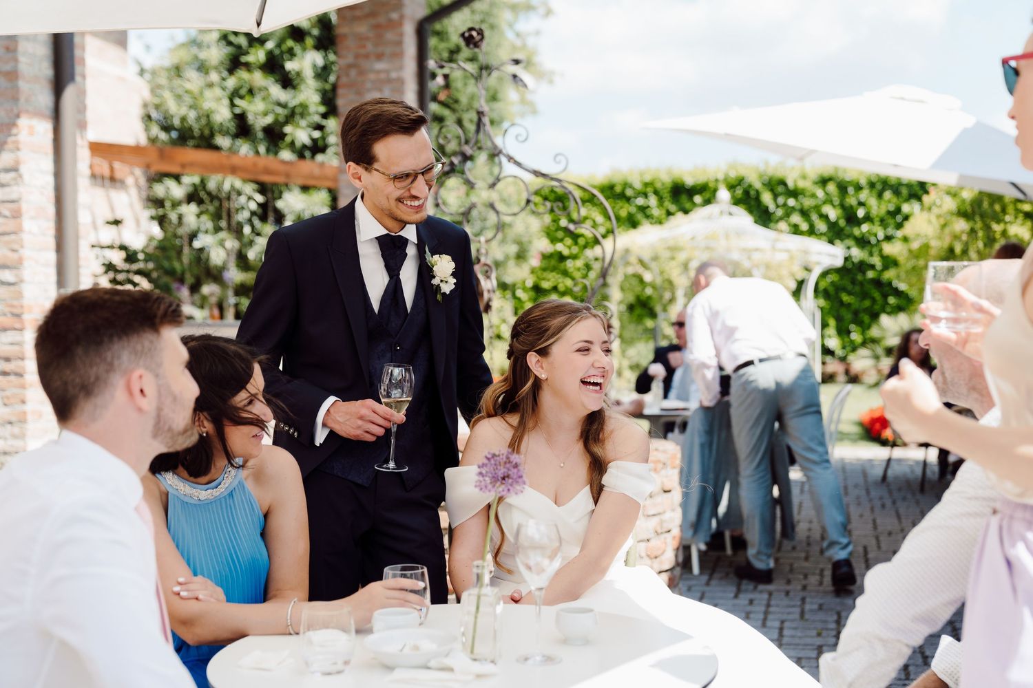 Groom in navy suit stands laughing with bride and guests at a sunny outdoor wedding reception table.