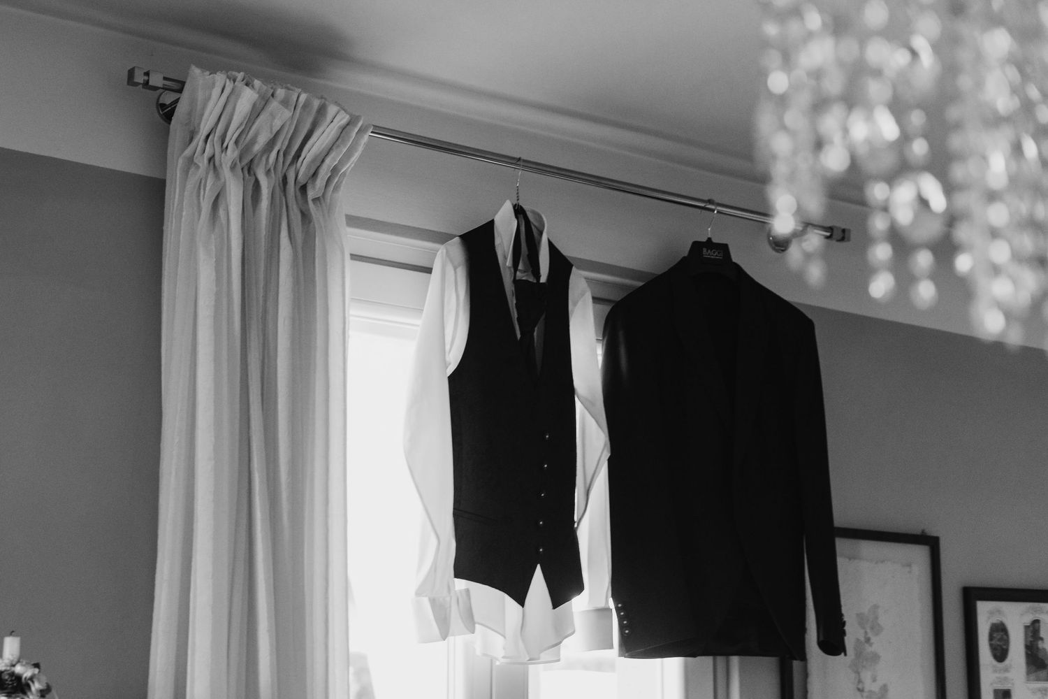 Black and white photo of a groom's suit vest and jacket hanging by a curtained window.
