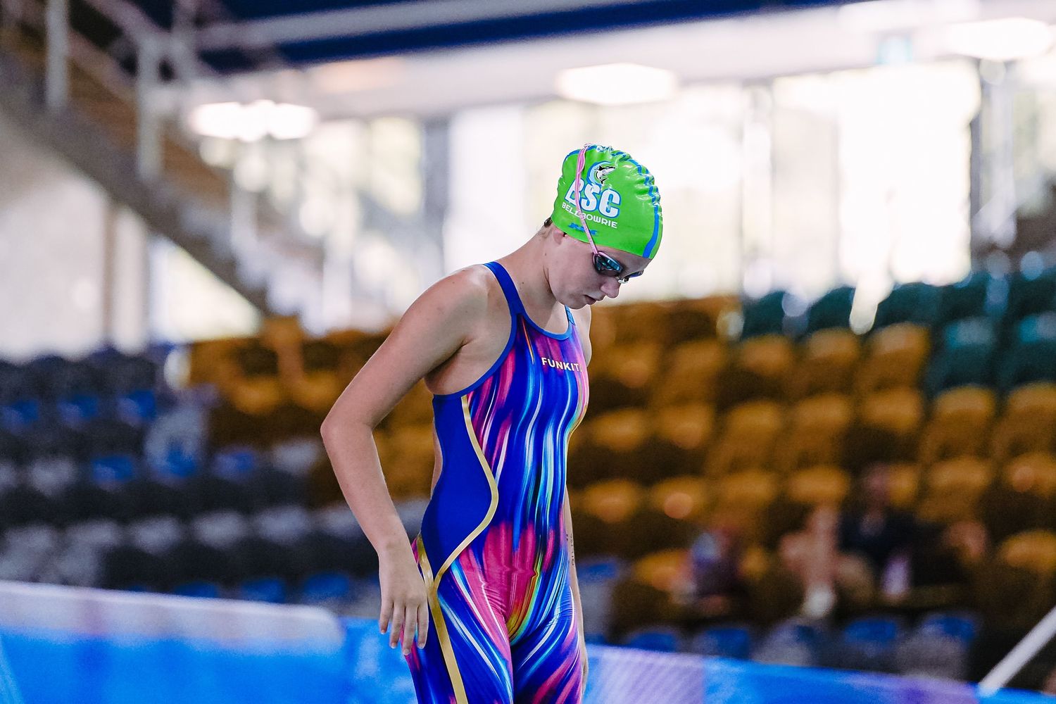 A swimmer competes in a pool lane during a competitive swimming race with timing boards visible in the background.