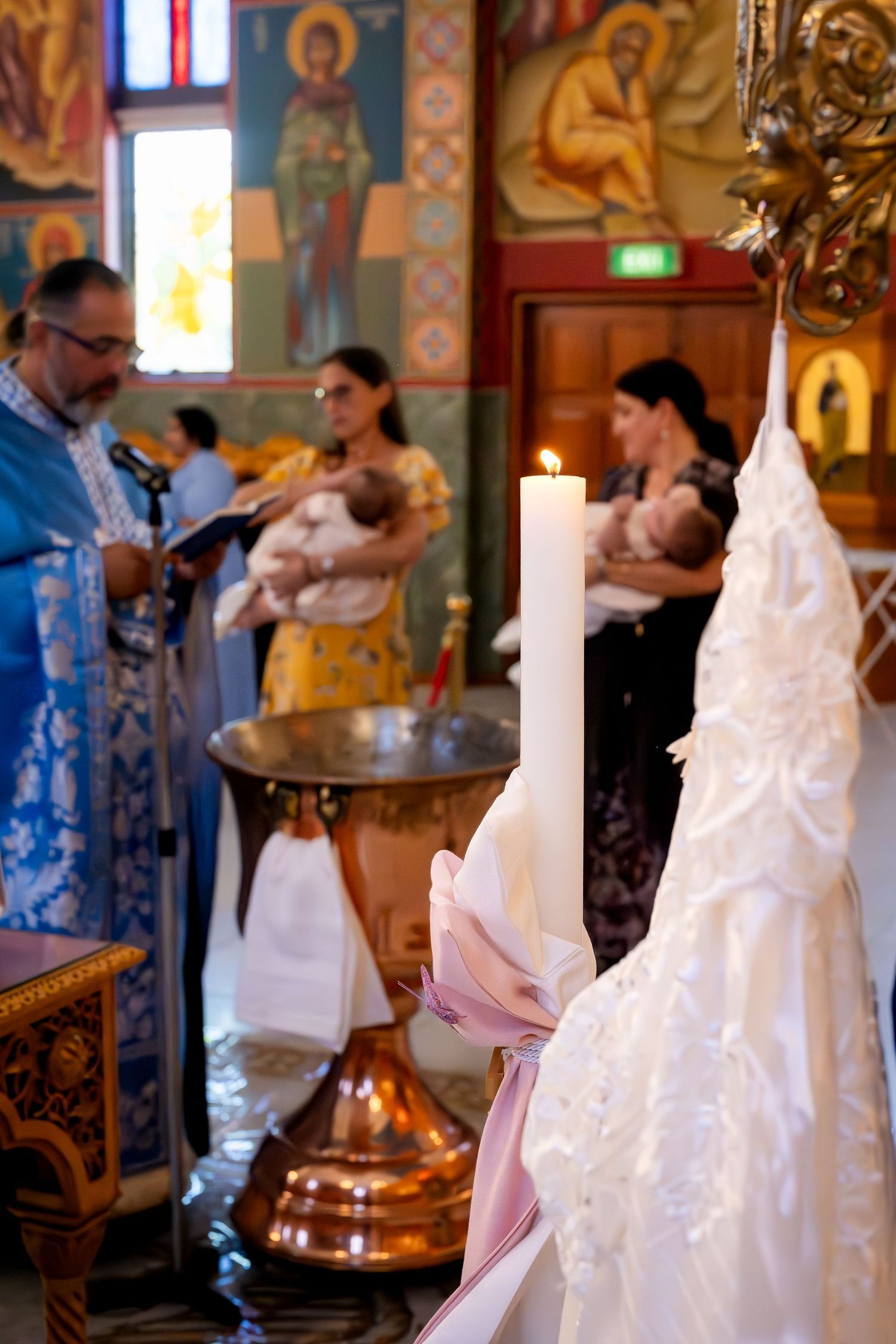 Orthodox Christian baptism ceremony with priest, two mothers holding infants near ornate copper font, white christening gown and candle.