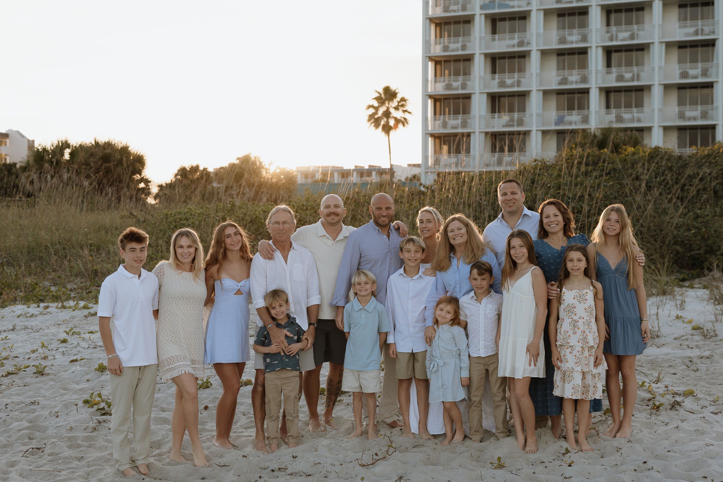 A group in coordinated neutral and blue outfits poses together on a sandy beach at sunset with the ocean behind them.