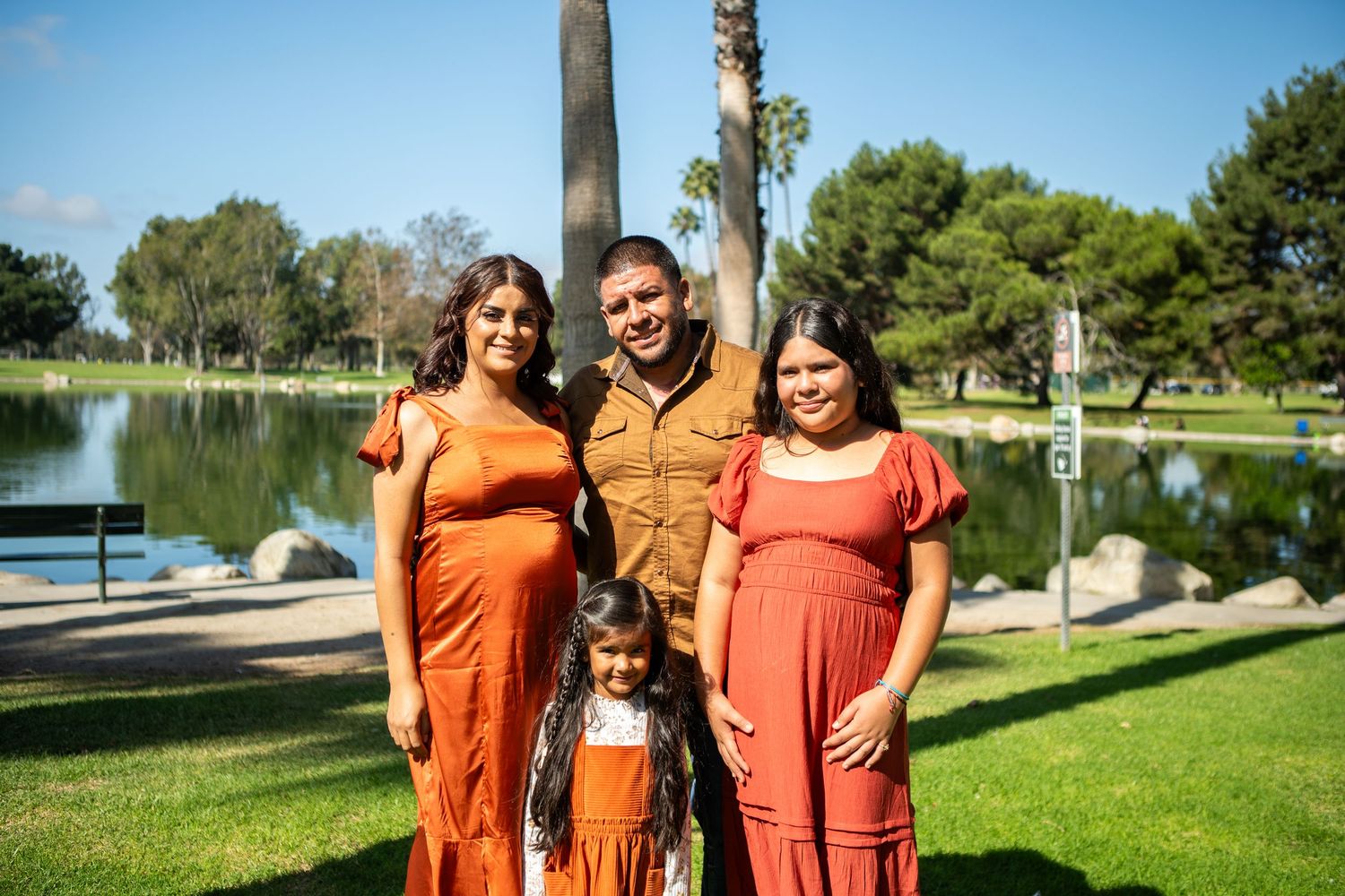 A group wearing matching orange outfits poses together in a sunny park setting with trees and a lake in the background.
