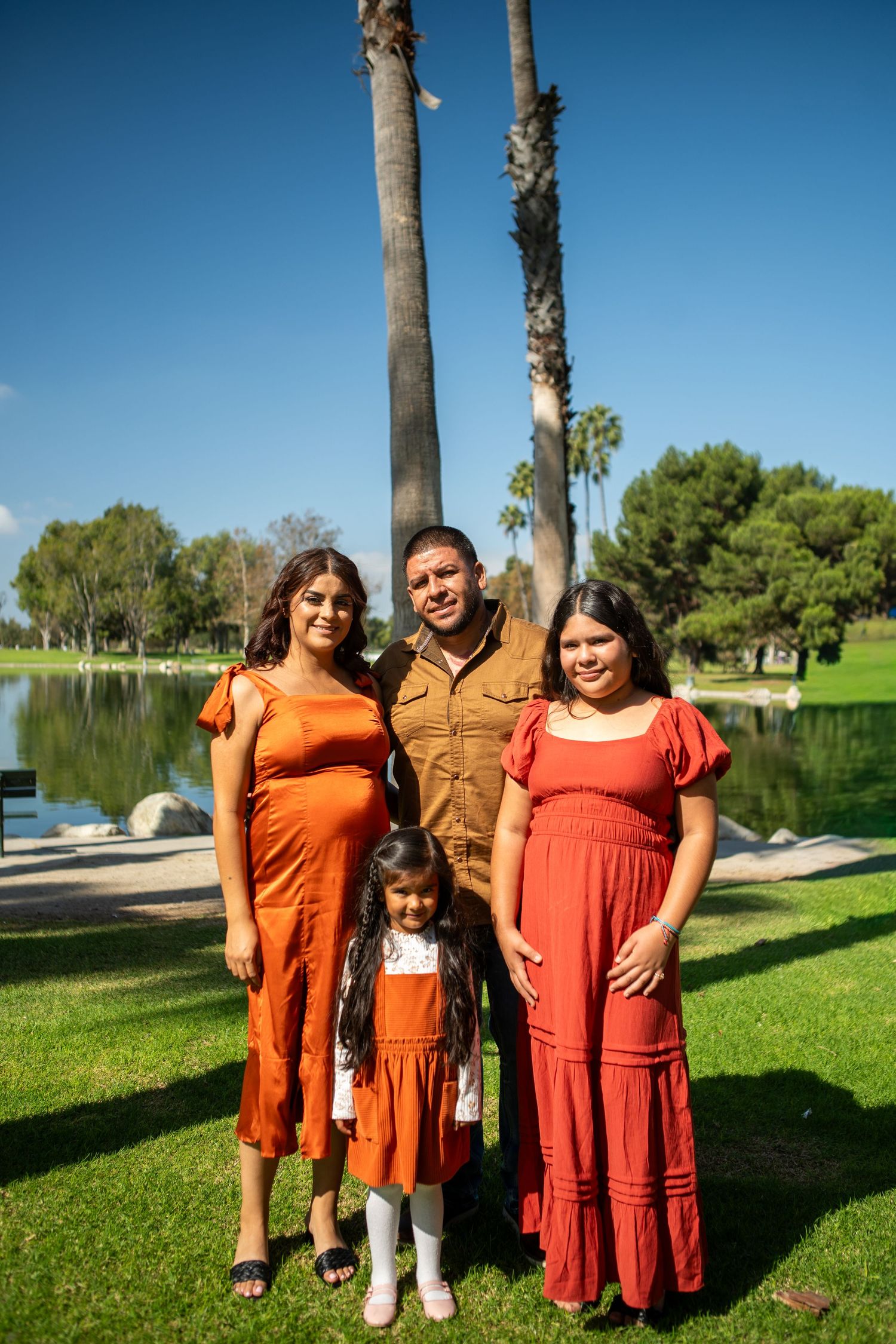 A group wearing matching orange and rust-colored dresses poses together on green grass near a lake on a sunny day.
