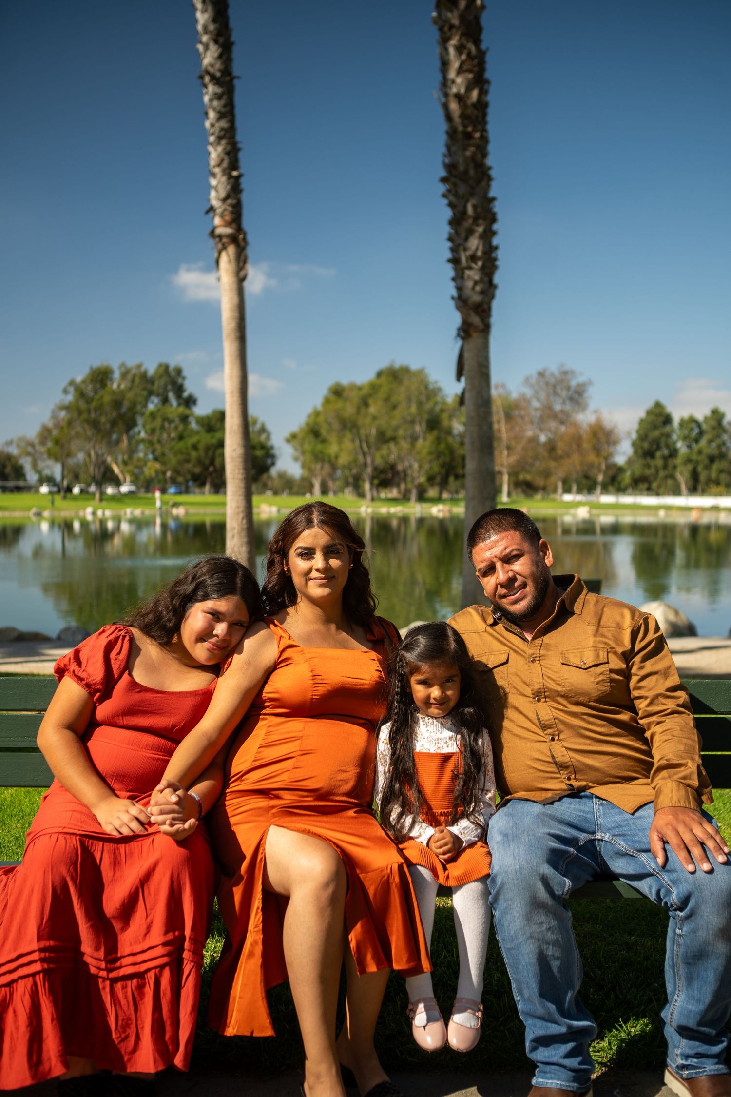 A family sits together on a bench near a peaceful lake surrounded by palm trees on a bright sunny day.