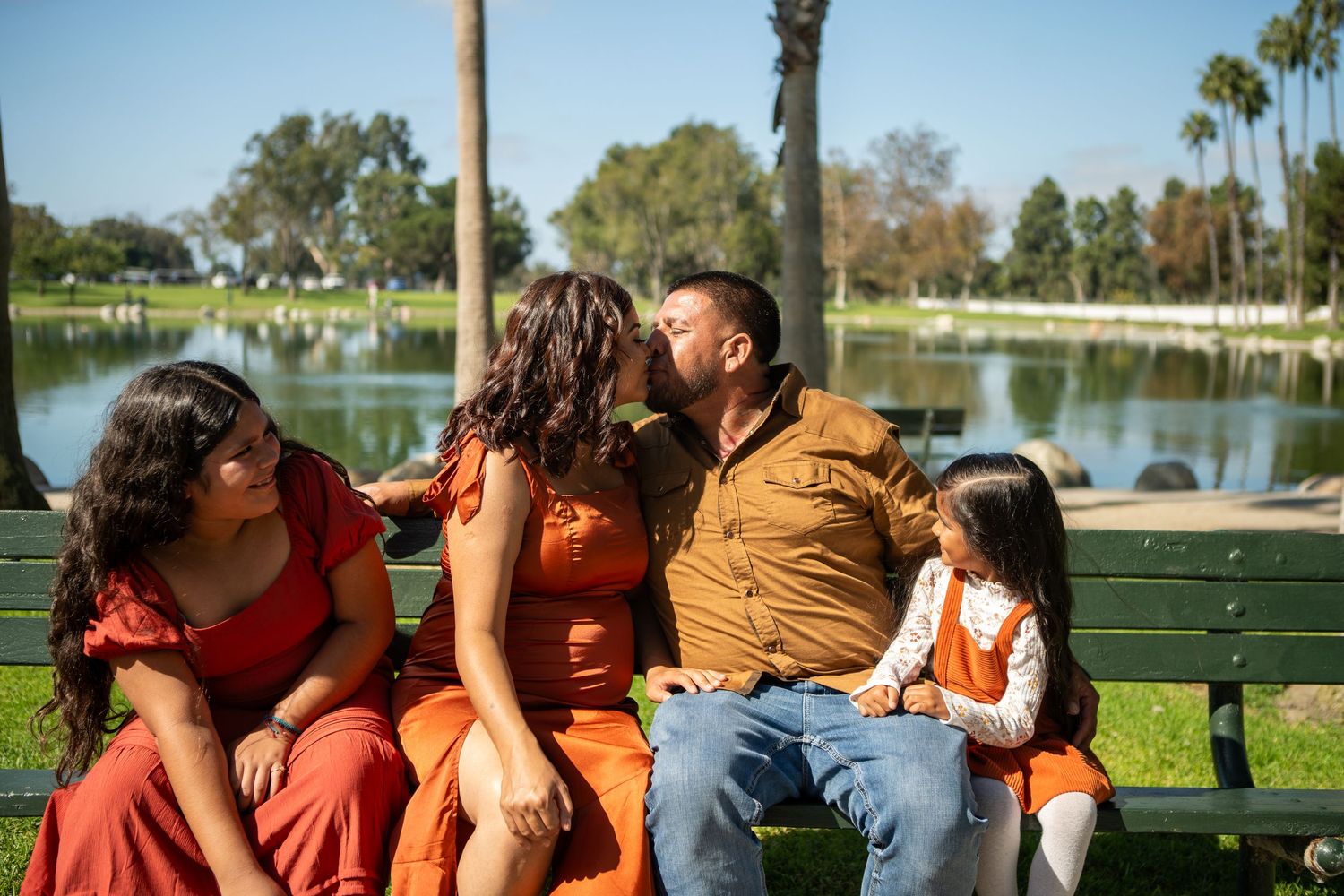 A group of people relaxing together on a bench by a lake in a park on a sunny day.