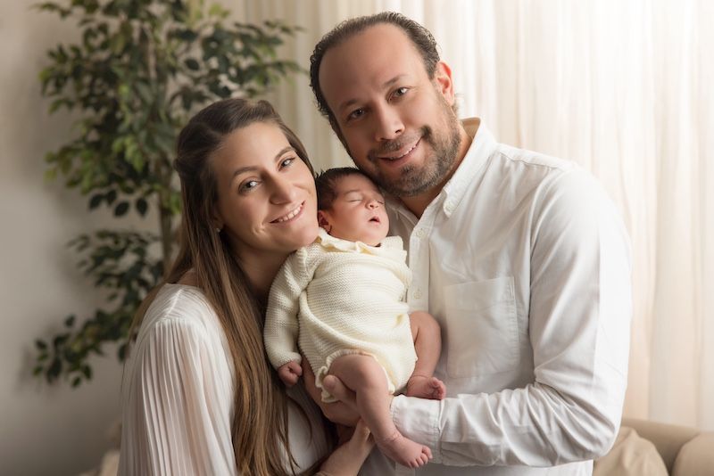 Happy couple in white clothing smiling while holding their newborn baby in a bright, cozy indoor setting.