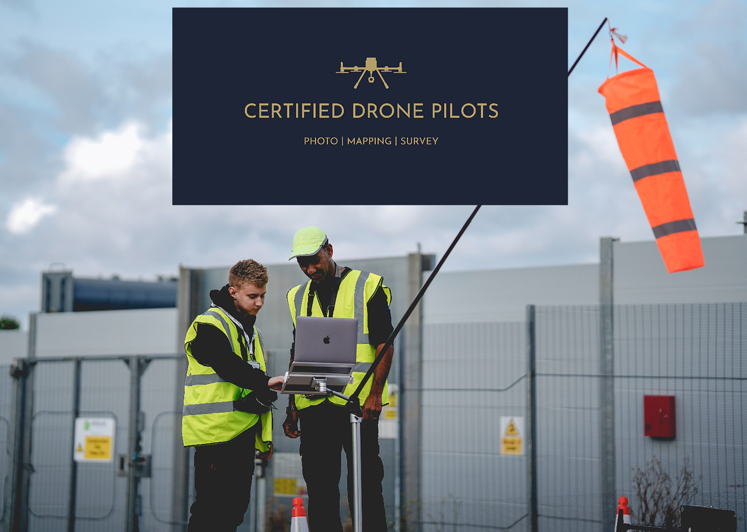 Two people in safety vests operate aerial site intelligence controls while monitoring a windsock at an industrial facility.