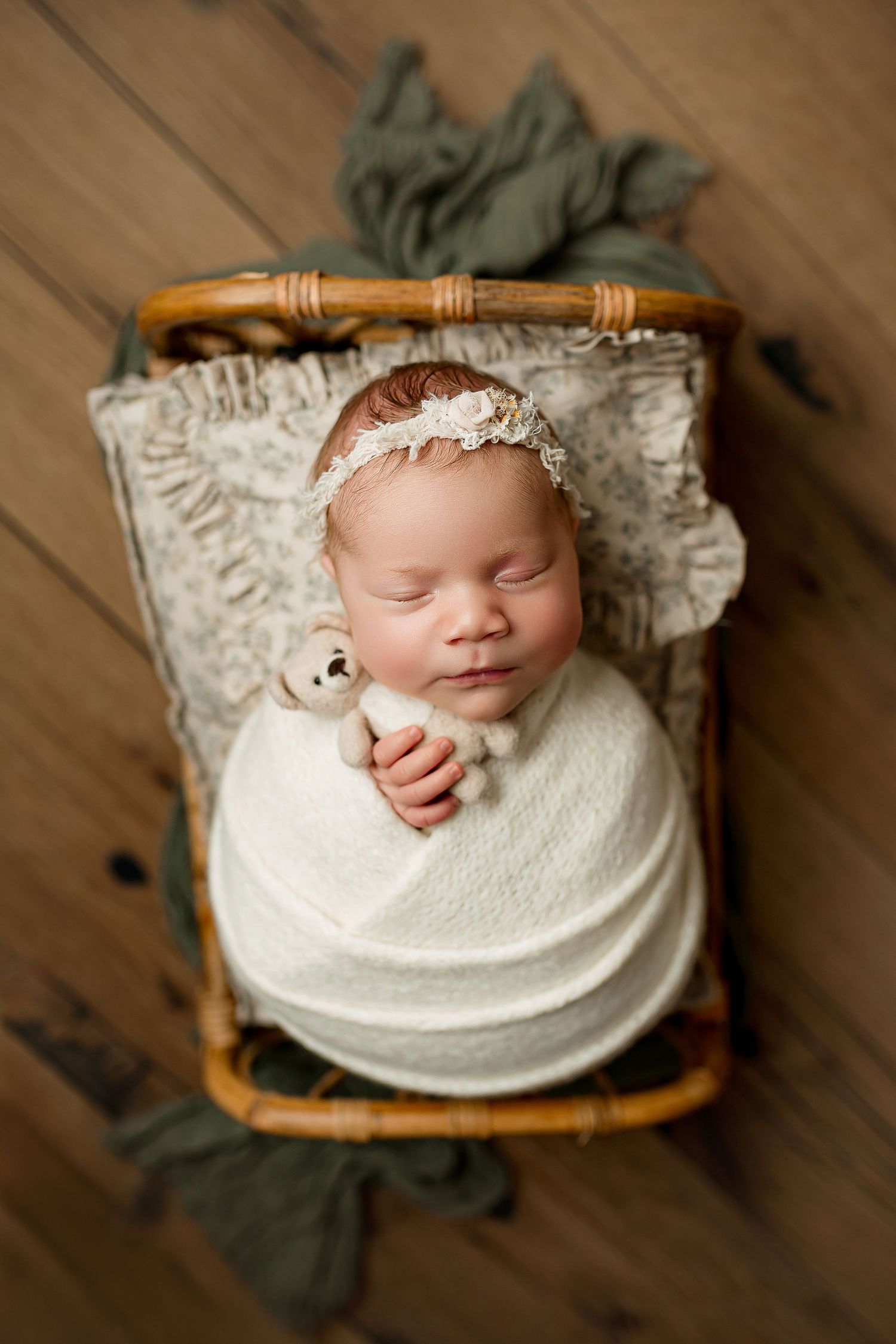 Newborn baby girl sleeping in rattan basket with sage green wrap and teddy bear on rustic wood floor at Summerville SC newborn studio