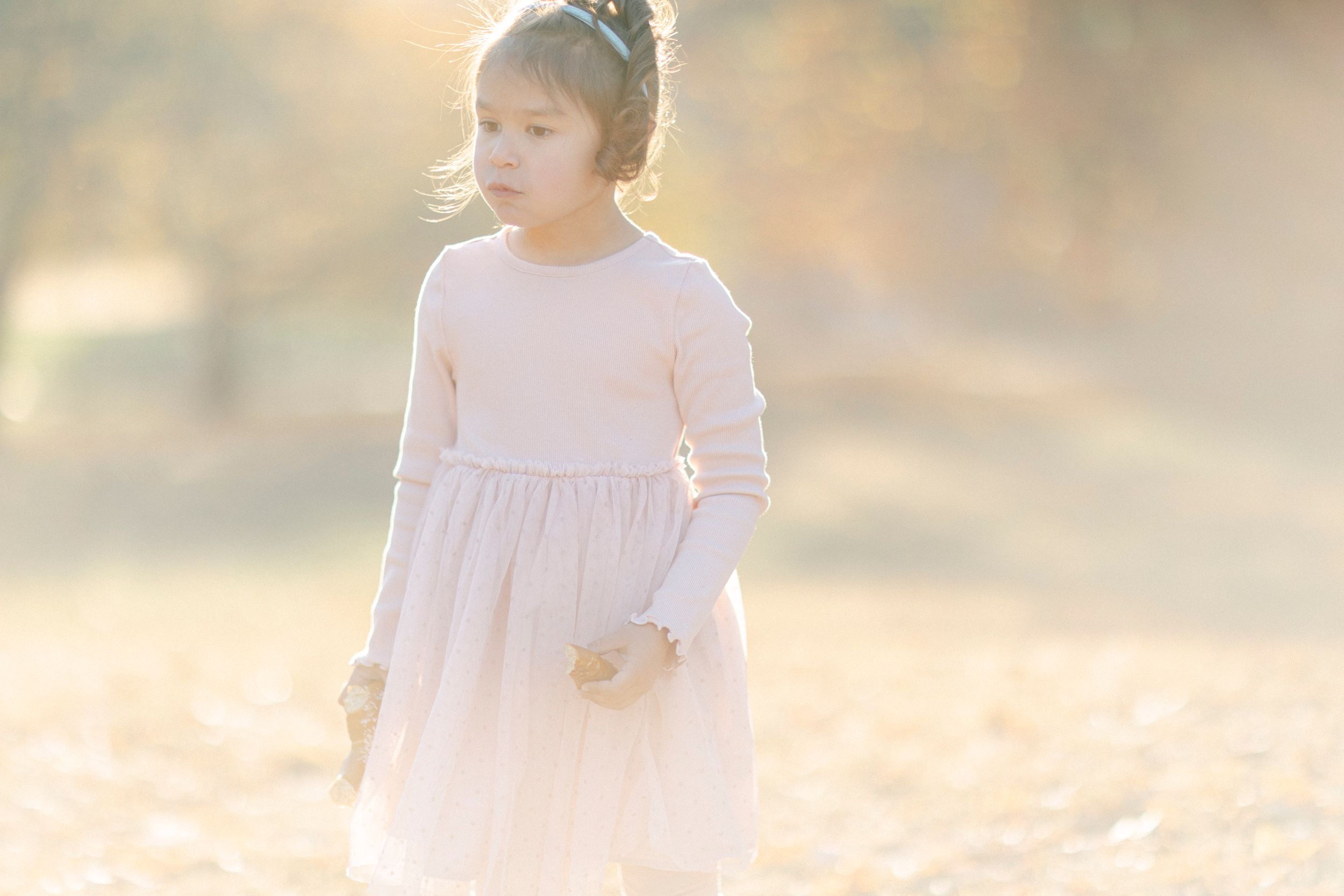 Child in white dress stands alone in dreamy golden sunlight outdoors.