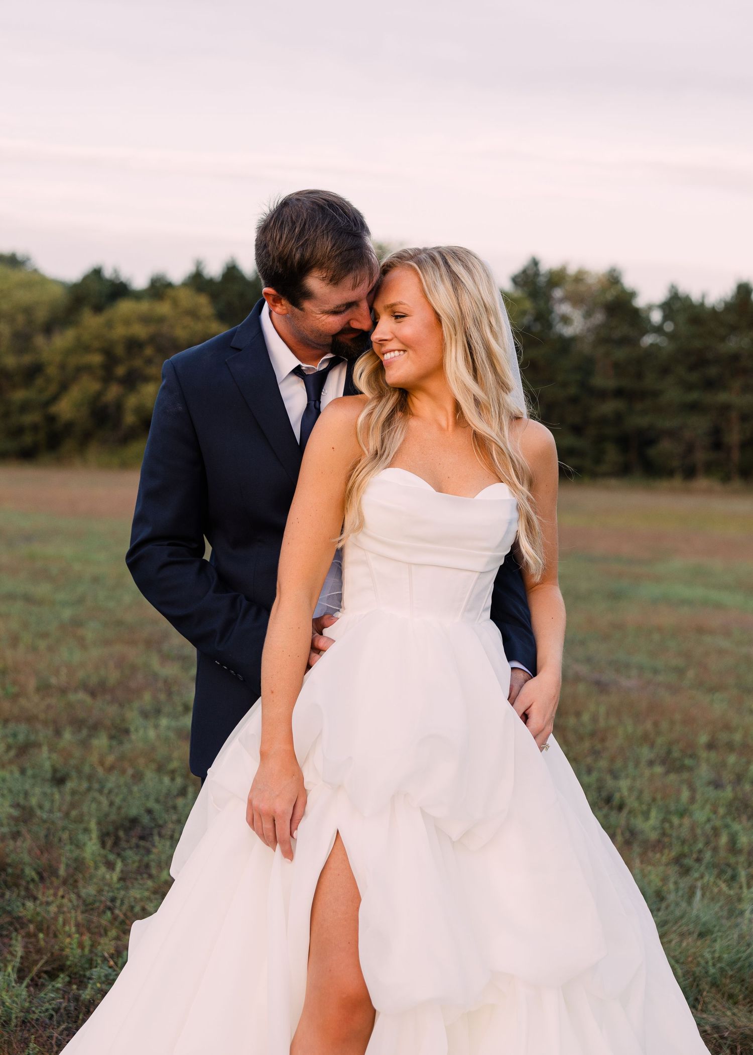 A smiling bride in a white strapless gown with a leg slit is embraced from behind by a groom in a navy suit in a grassy field.