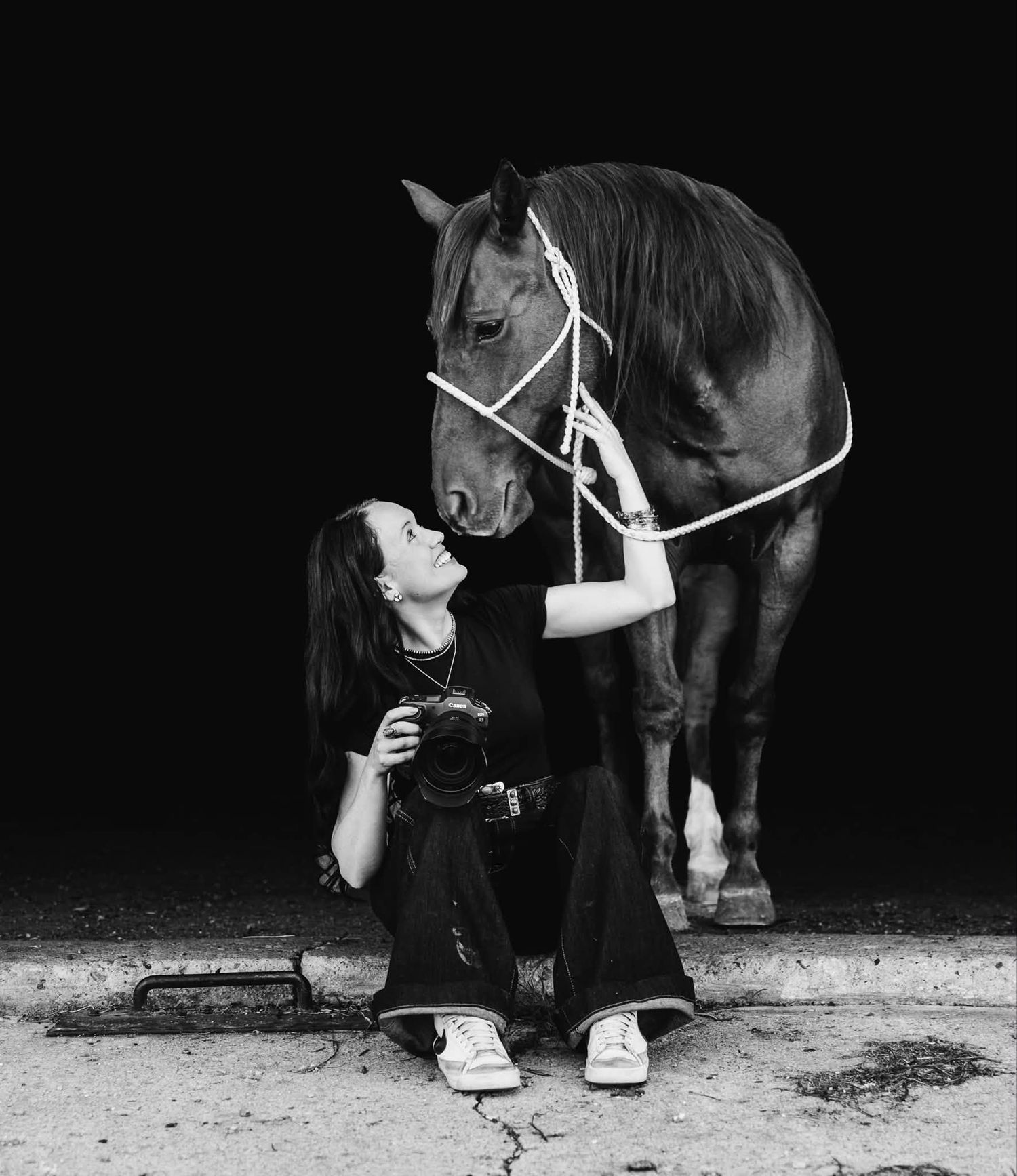 Woman in black clothes sits on the ground holding a camera and gazes up at a dark horse in a rope halter in black and white.