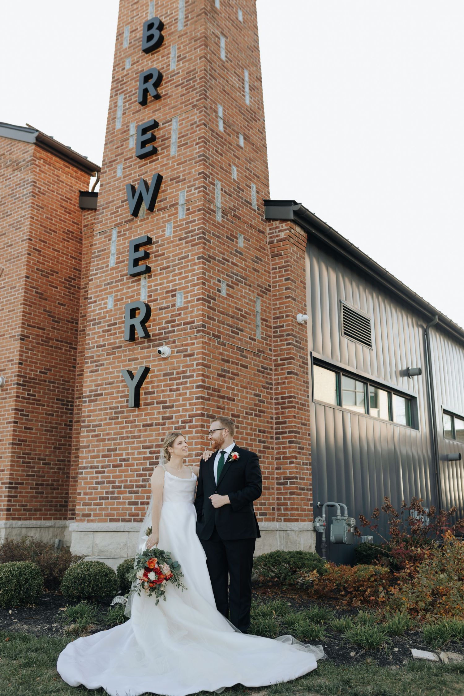 Wedding couple poses in front of a tall brick brewery building with vintage industrial architecture.