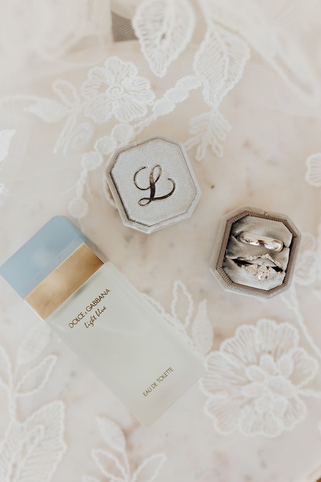 Wedding rings displayed in a gray velvet box with monogram lid and light blue perfume bottle on delicate lace fabric.