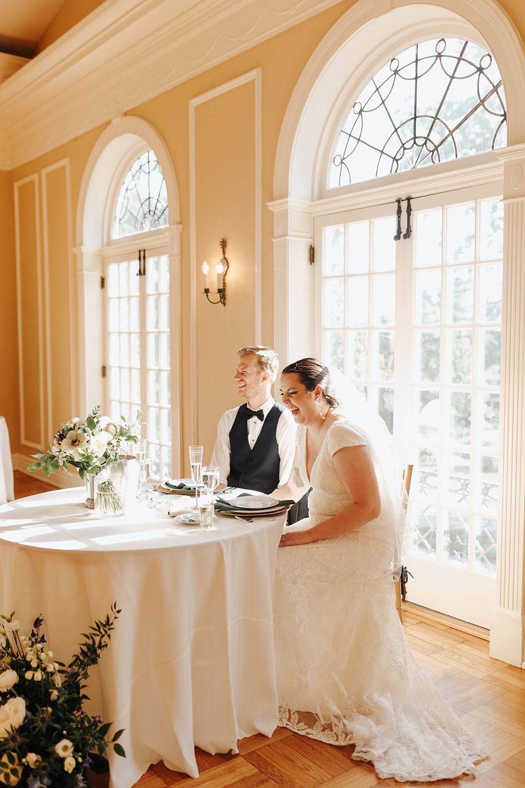 Elegant wedding reception setup in a bright room with arched windows, white tablecloth and floral arrangements.