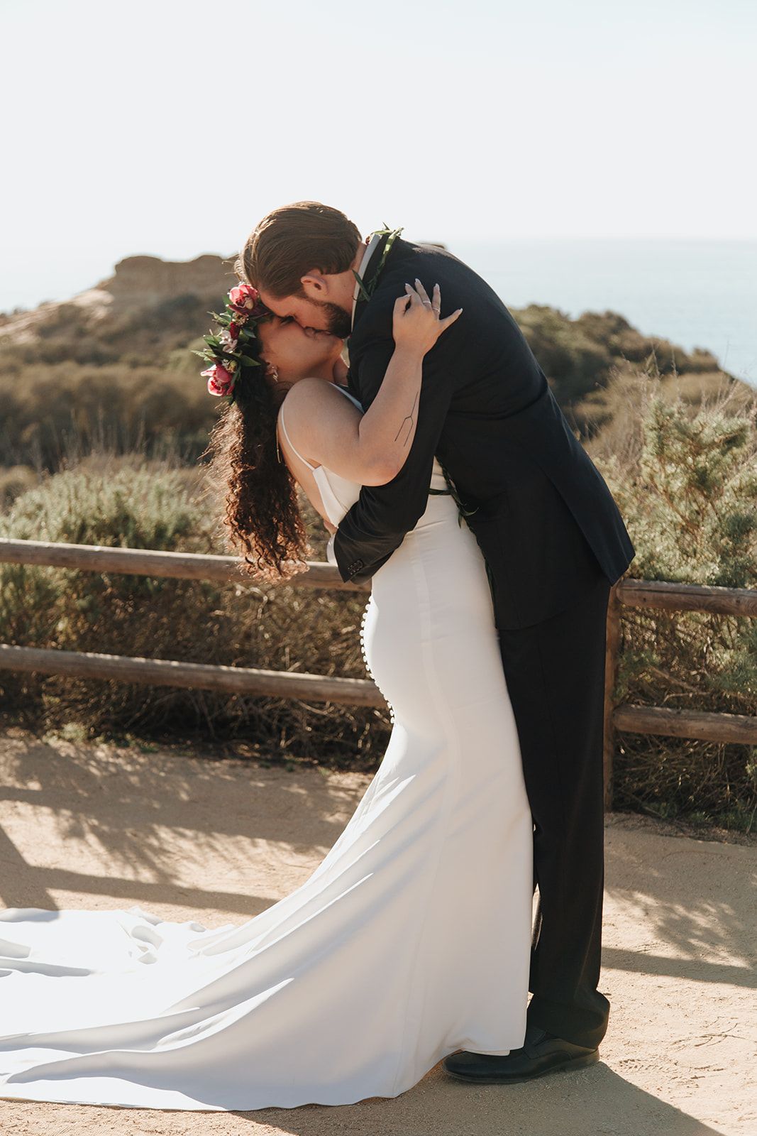 A bride and groom share a romantic kiss on a scenic coastal clifftop with flowing white dress and floral crown at sunset.