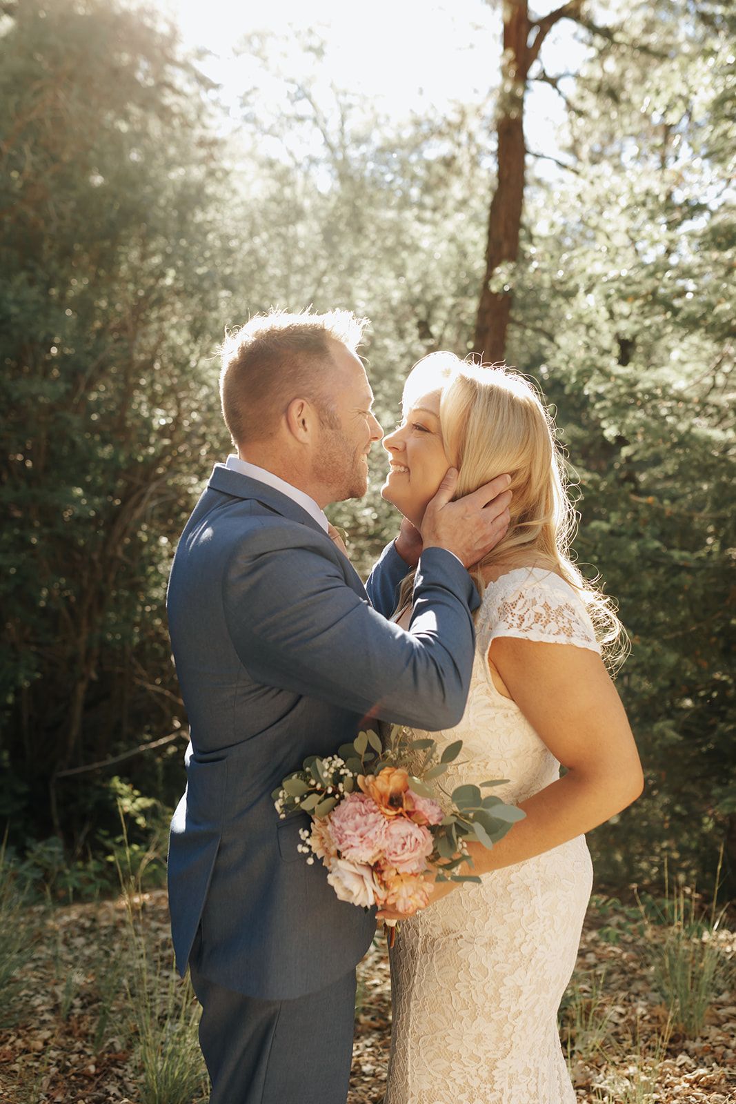 Romantic wedding couple shares tender moment in sunlit forest while bride holds pink and peach floral bouquet.