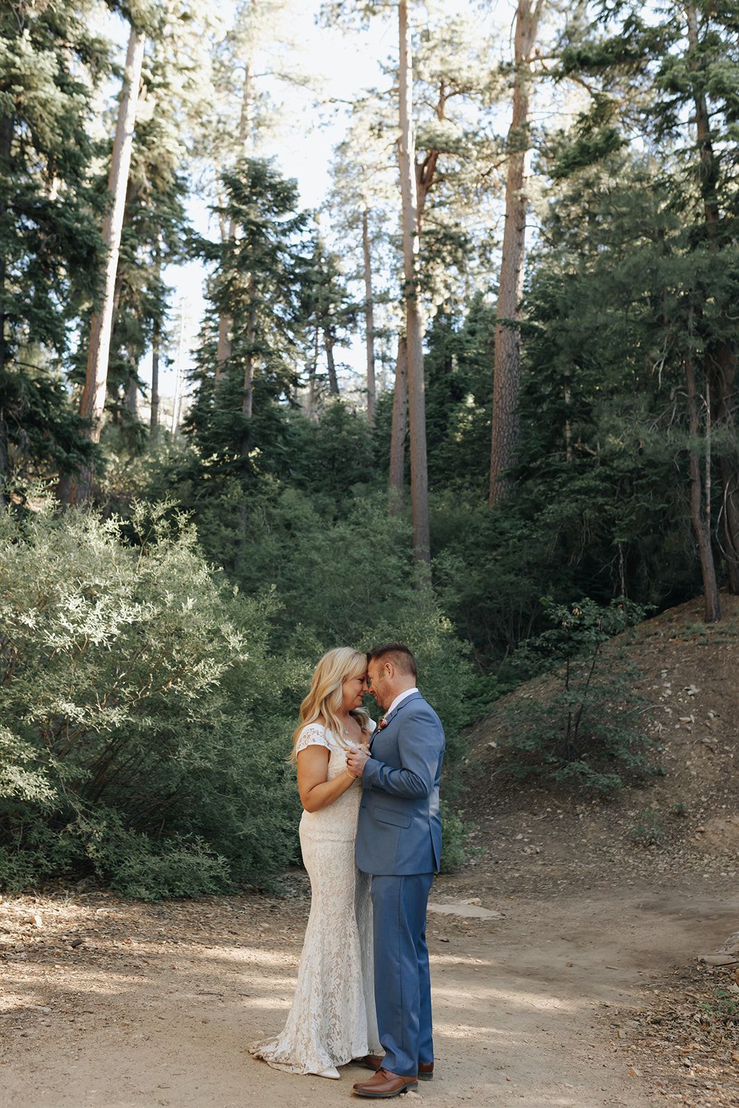 A couple shares an intimate moment on a forest trail surrounded by tall evergreen trees on their wedding day.