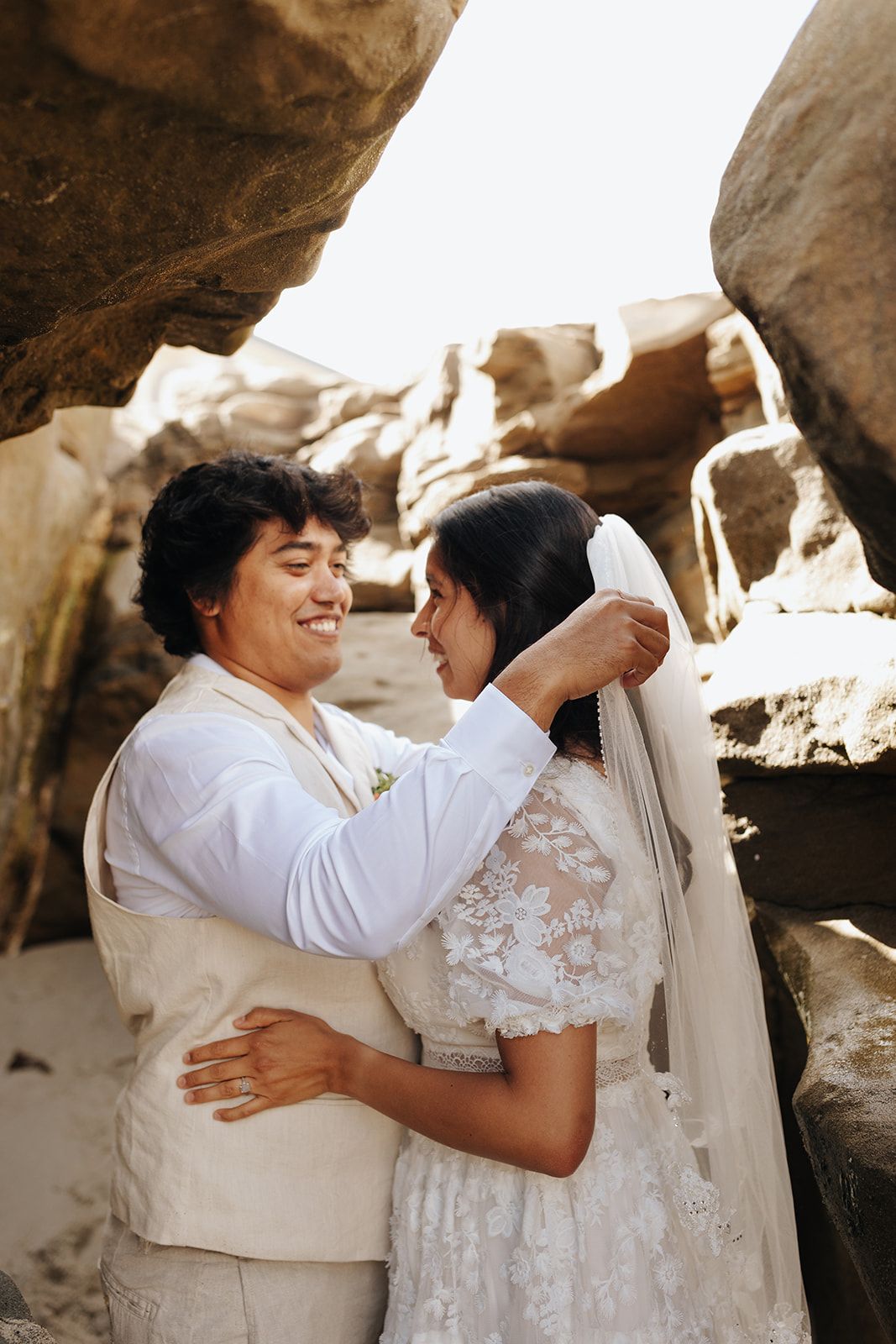 A couple shares a tender moment on their wedding day among coastal rock formations, wearing white attire.