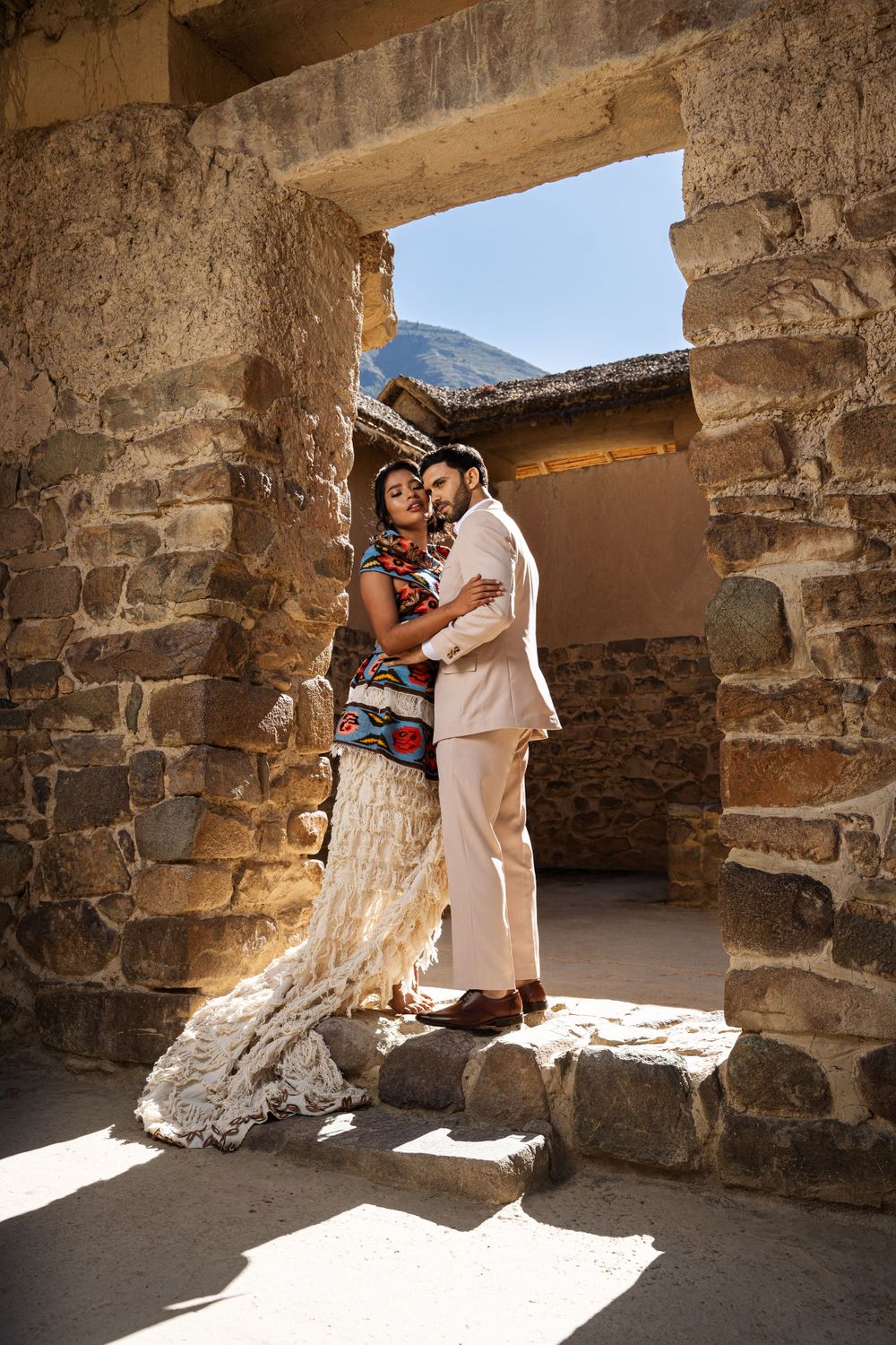 A couple in elegant attire posing in an ancient stone doorway with mountains visible beyond.
