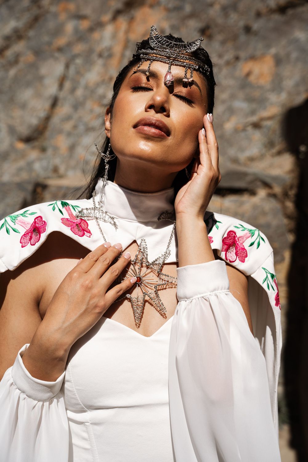 Model wearing white dress with floral embroidery and ornate headpiece poses against stone wall.