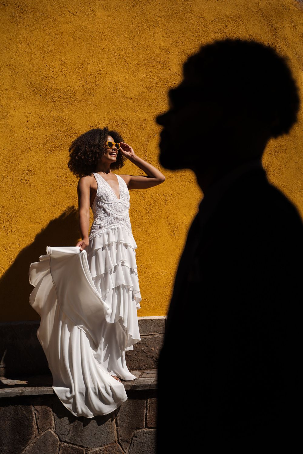 A bride in a flowing white tiered dress poses against a yellow wall casting a dramatic shadow.