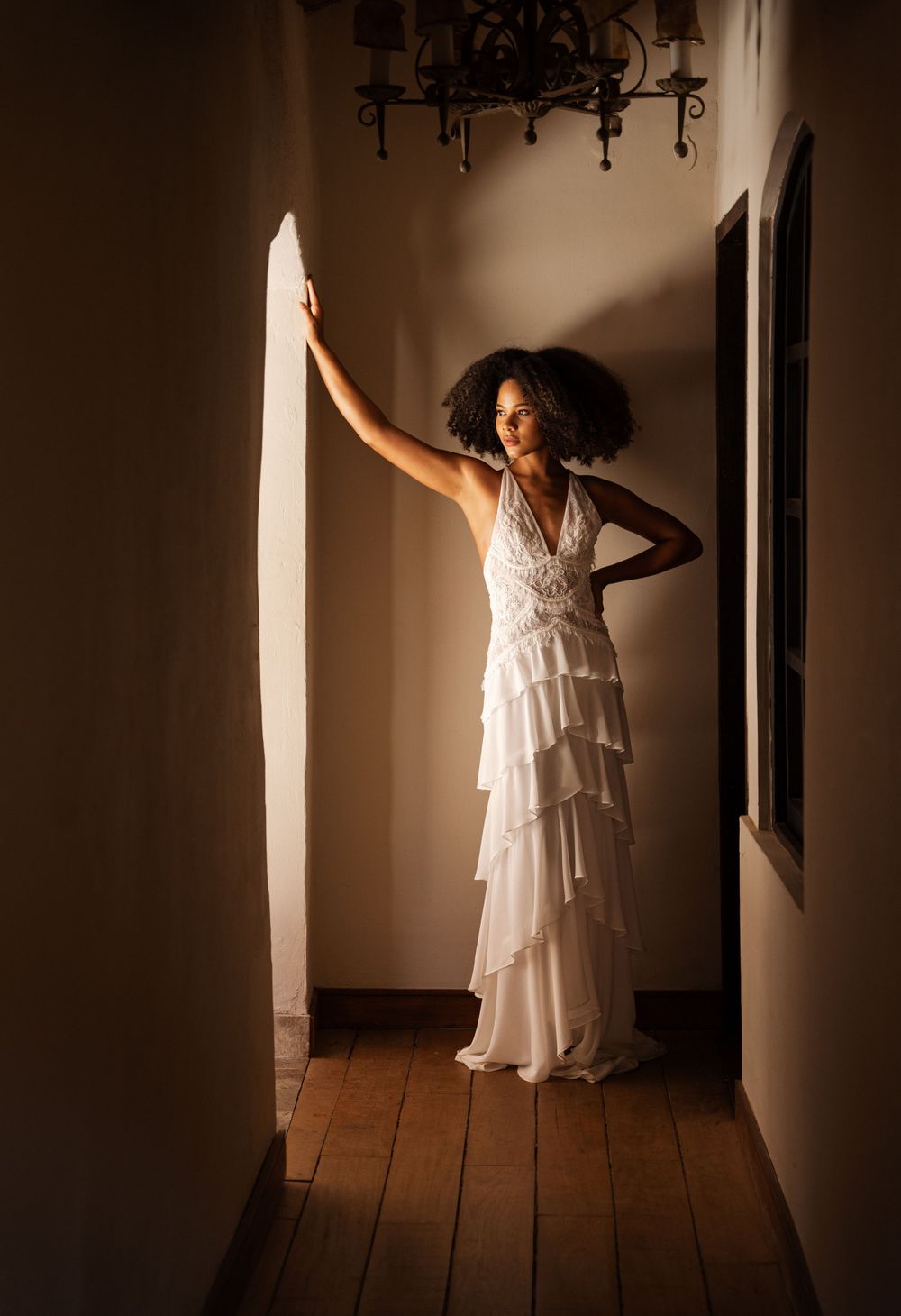 A bride in a white ruffled gown stands in a sunlit hallway near an arched doorway.