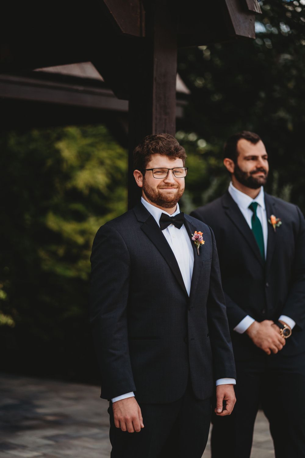 Two men in formal tuxedos stand together at an outdoor wedding venue.