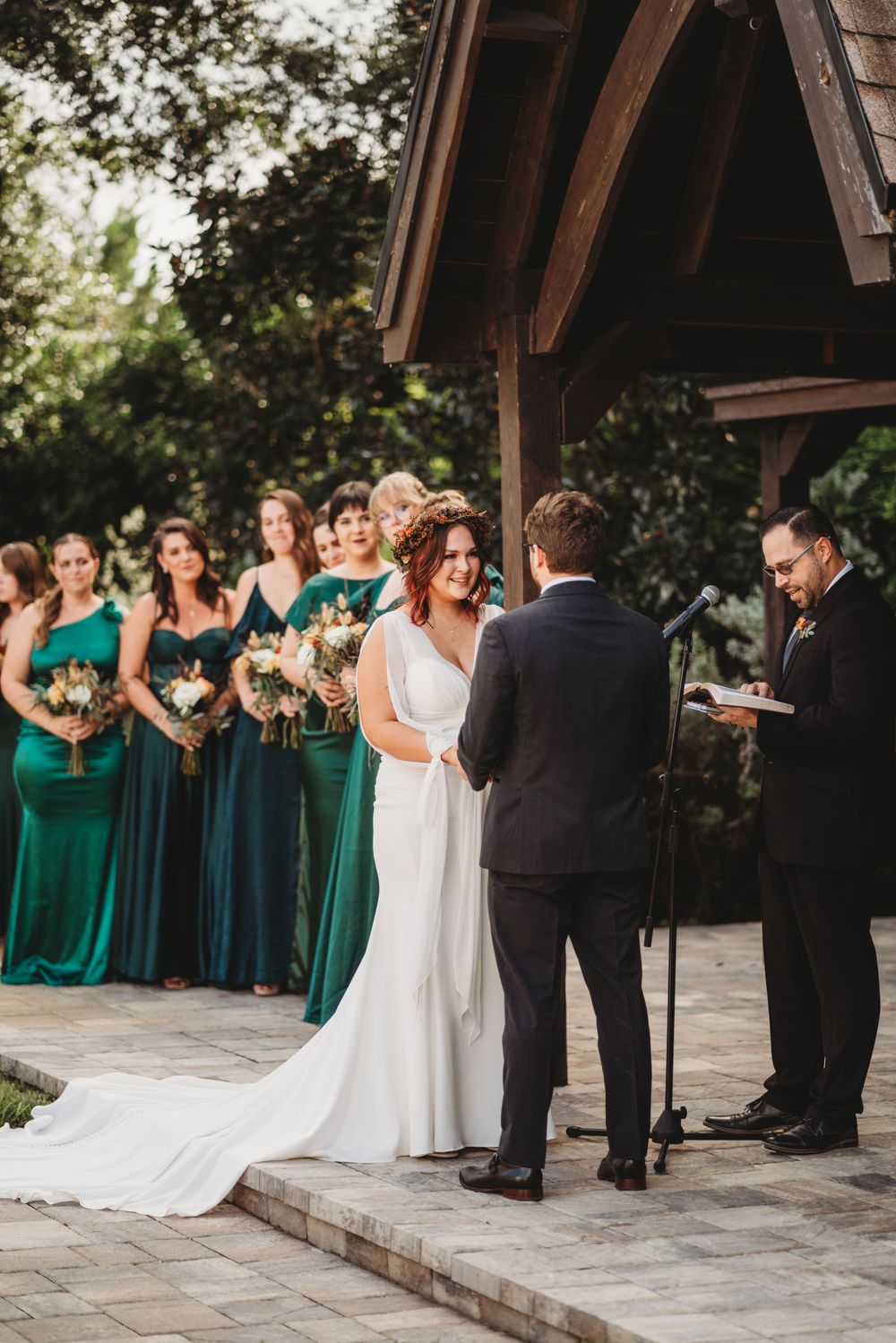 A wedding ceremony with bridesmaids in emerald green dresses under a wooden pavilion.