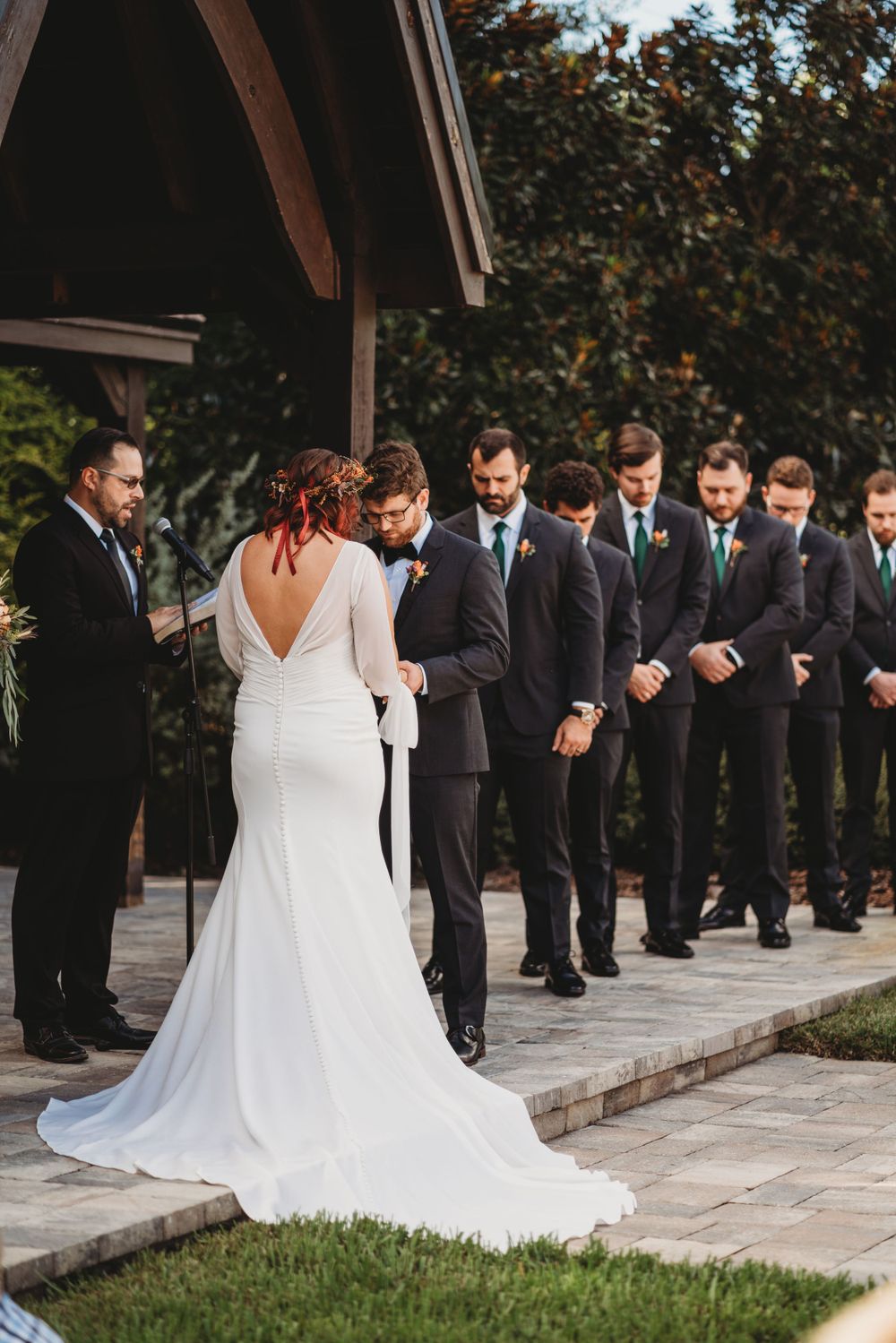 A bride in a white dress stands before a line of groomsmen at an outdoor ceremony.