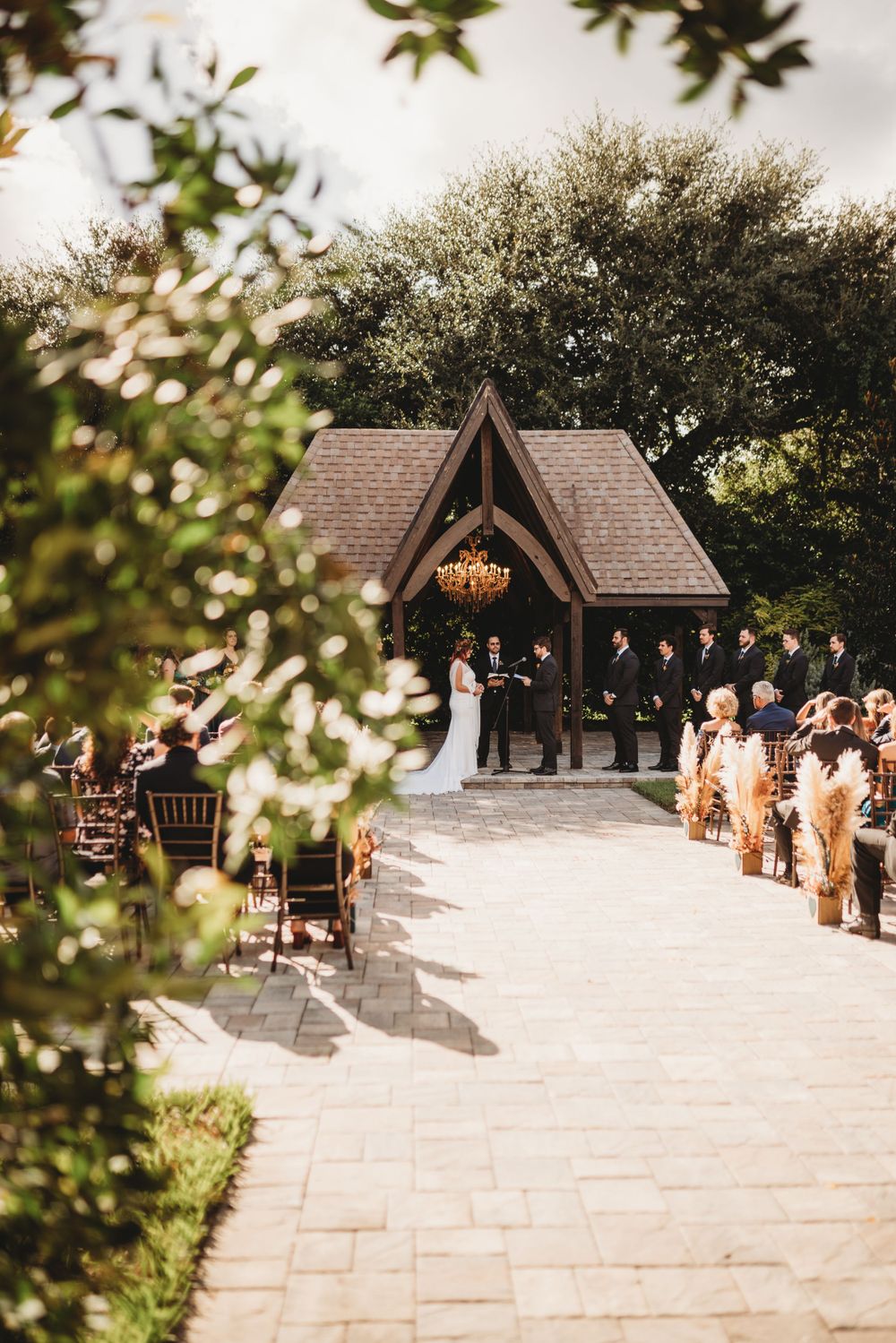A rustic wooden chapel frames a wedding ceremony amid lush greenery.