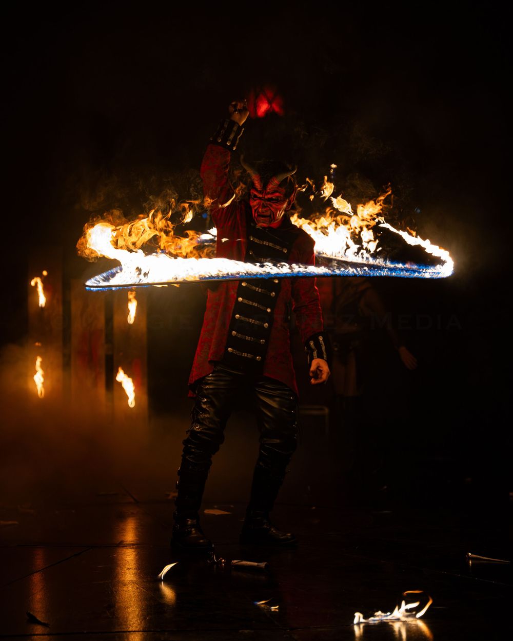 Performer spins flaming hoop in dramatic dark stage performance.