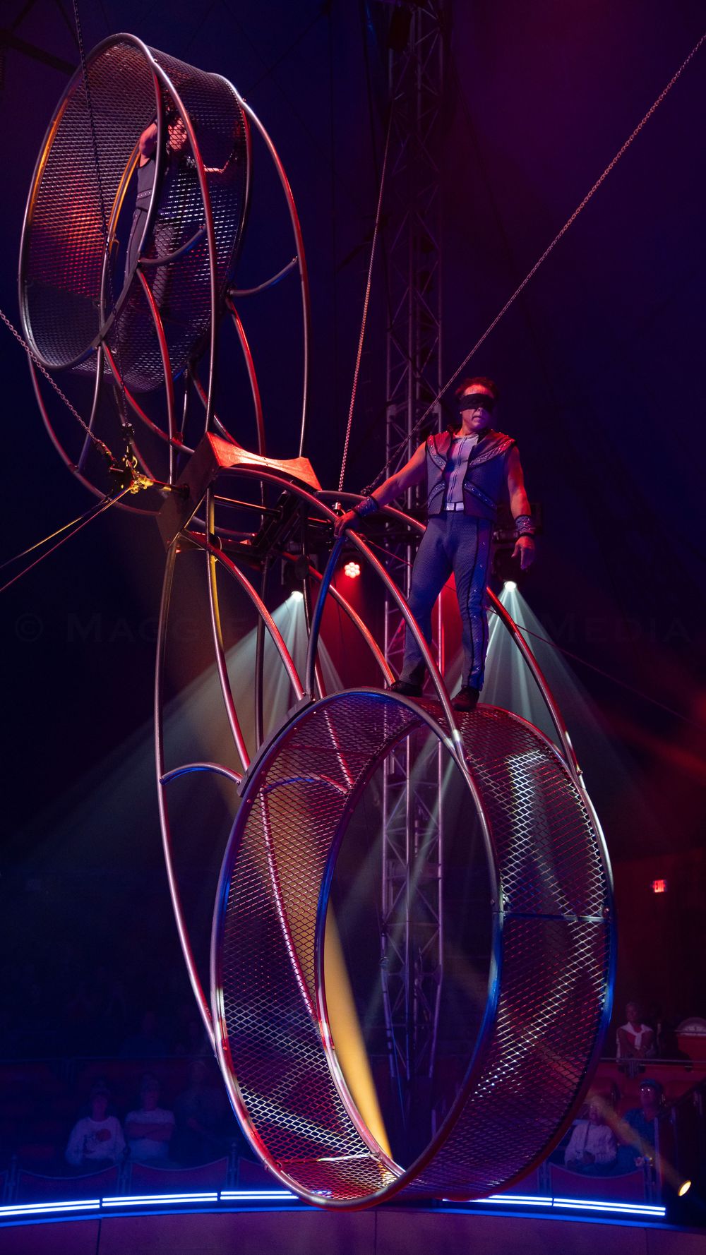 Acrobat Noe Espana performs on large metal wheel apparatus under dramatic stage lighting.